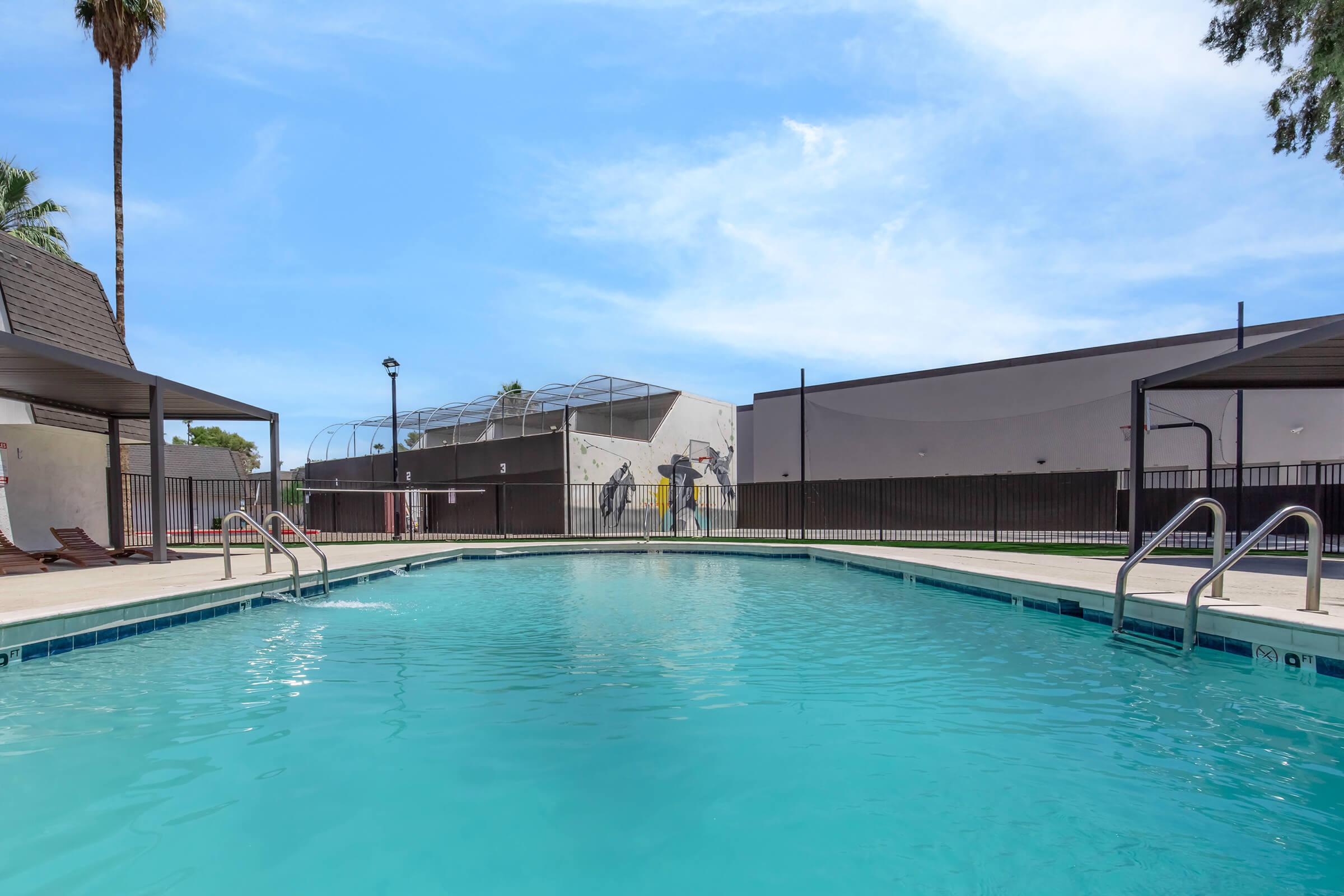 A clear blue swimming pool surrounded by a patio area with lounge chairs. In the background, there are buildings with a fence, and a construction area visible. The sky is bright with a few clouds, creating a sunny atmosphere.