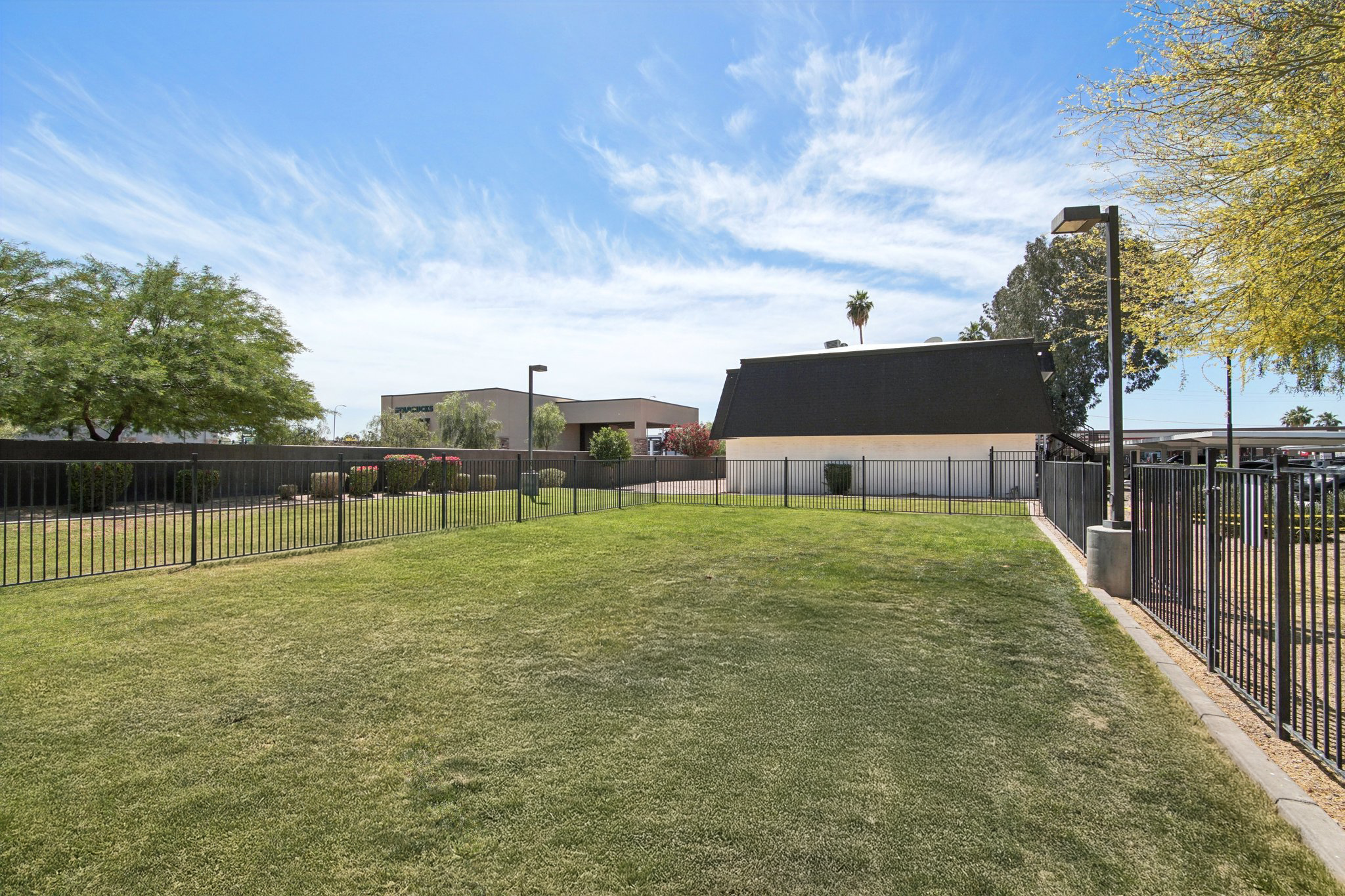 Open grassy area enclosed by a black fence, with shrubs and trees in the background. A building with a peaked roof is visible behind the greenery. The sky is bright blue with wispy white clouds. The scene conveys a sunny outdoor space suitable for recreational activities.