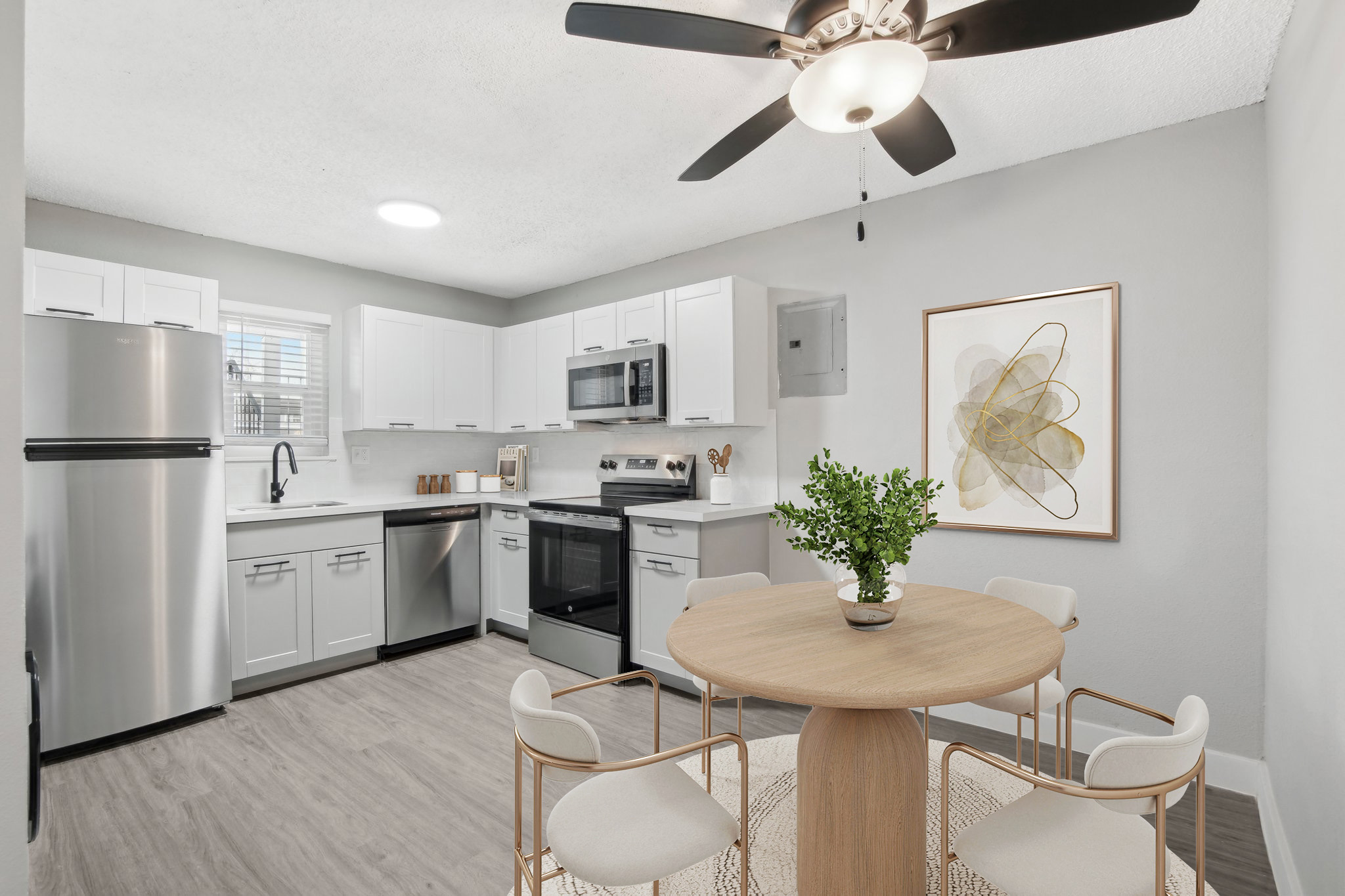 A modern kitchen with white cabinetry and stainless steel appliances. Features include a round wooden dining table with four white chairs, a ceiling fan, and a decorative plant. A piece of abstract art hangs on the wall, and natural light floods the space through a window.
