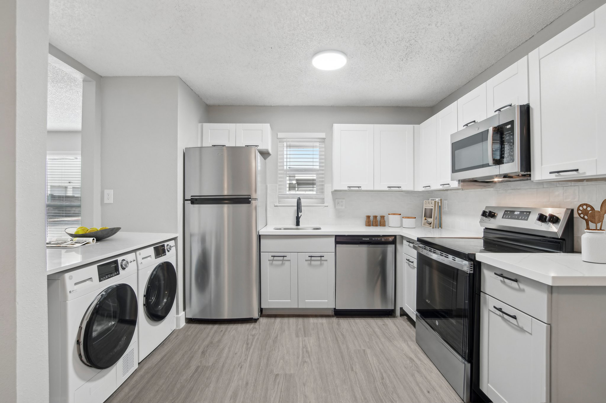 A modern kitchen featuring white cabinetry, stainless steel appliances including a refrigerator, stove, and dishwasher. There's an integrated laundry area with front-loading washers. The countertop is light-colored, and a window provides natural light. The floor is wood-like laminate.