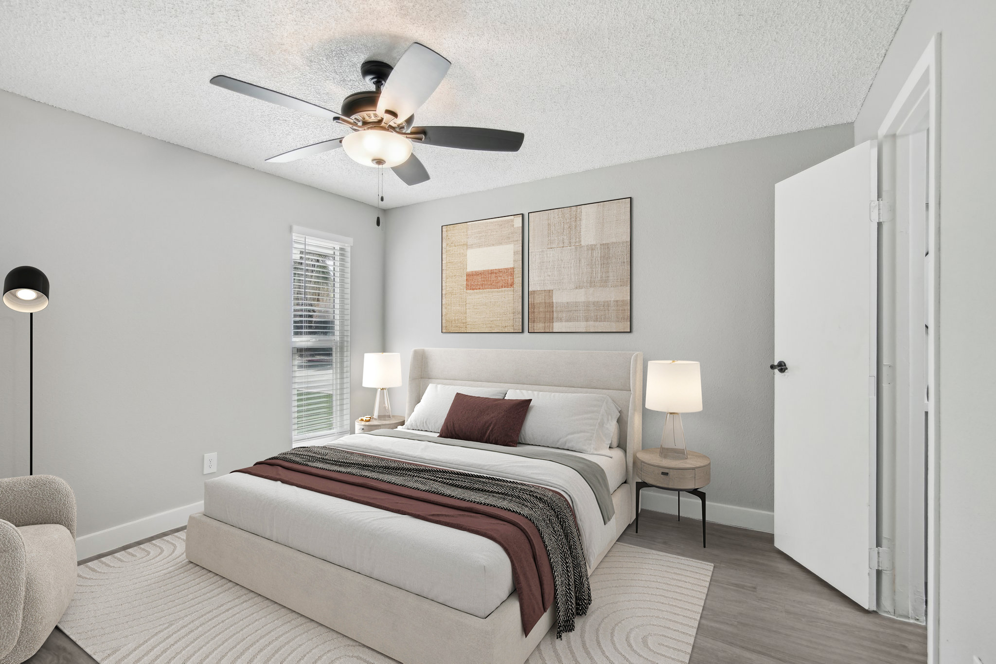 A modern bedroom featuring a light-colored bed with decorative throw pillows, a ceiling fan overhead, and two bedside lamps. There are abstract wall art pieces above the bed, a cozy area rug on the floor, and a light gray wall. A door is visible on the right, and a soft armchair is in the corner.