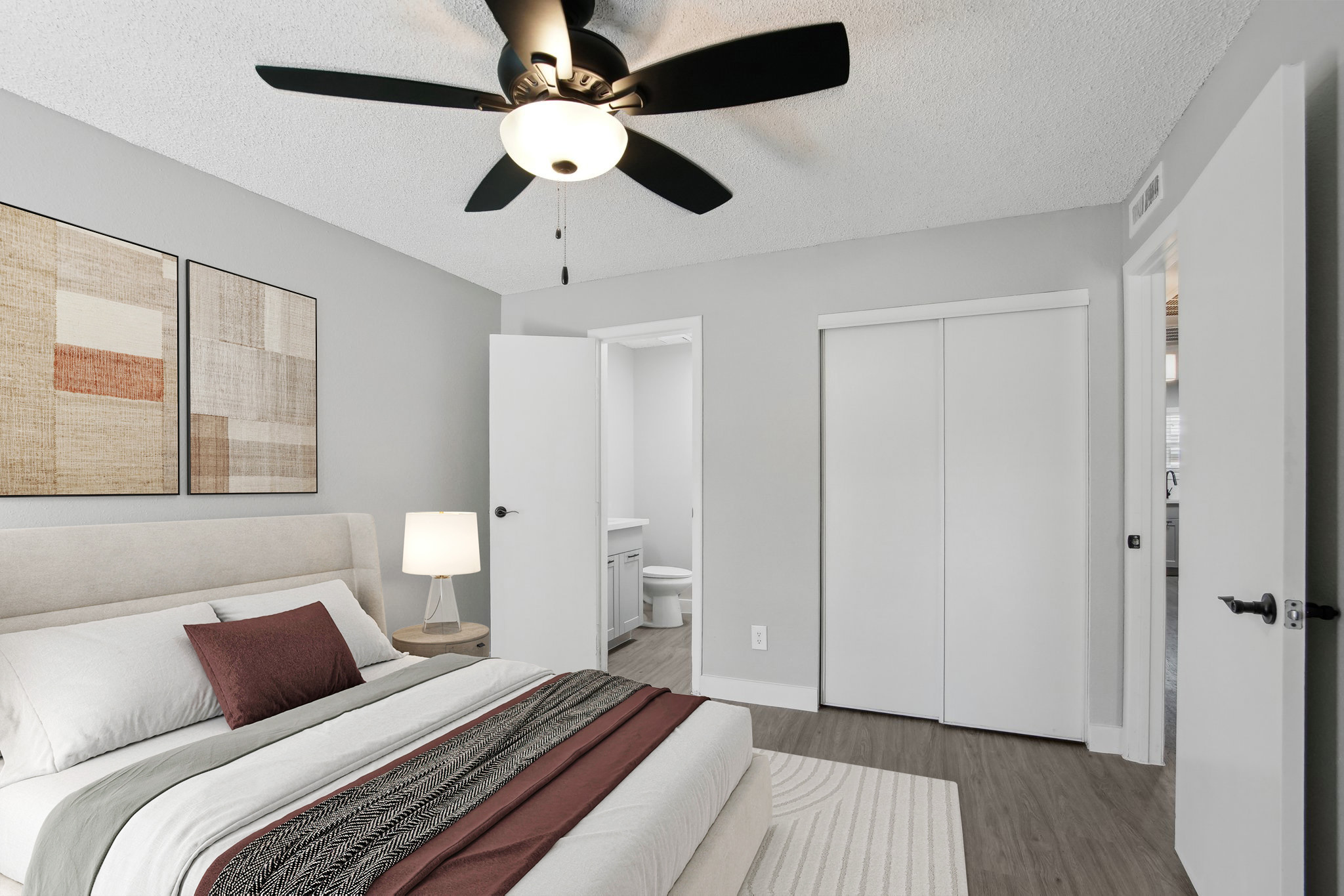 Cozy bedroom featuring a bed with decorative pillows and a striped blanket, a bedside lamp, and a ceiling fan. Light gray walls and contemporary artwork adorn the space. The room has a closet with sliding doors and an open door leading to a bathroom, creating a modern and inviting atmosphere.