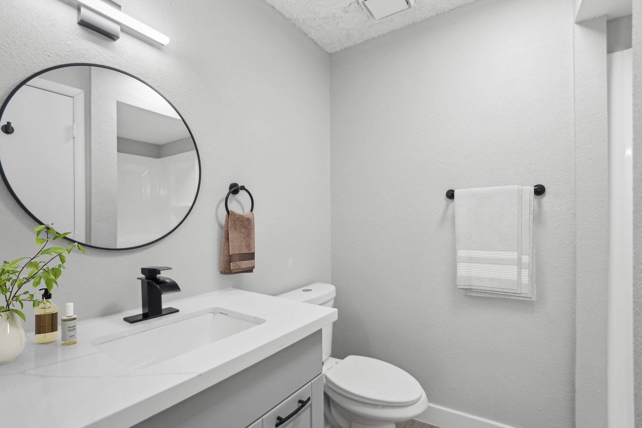 Modern bathroom with light gray walls, a round mirror above a white sink with a black faucet. A small plant and toiletries are on the countertop. There's a towel rack with a brown towel and a white towel on a hook nearby. A toilet is visible, against the wall, contributing to a clean and minimalistic design.