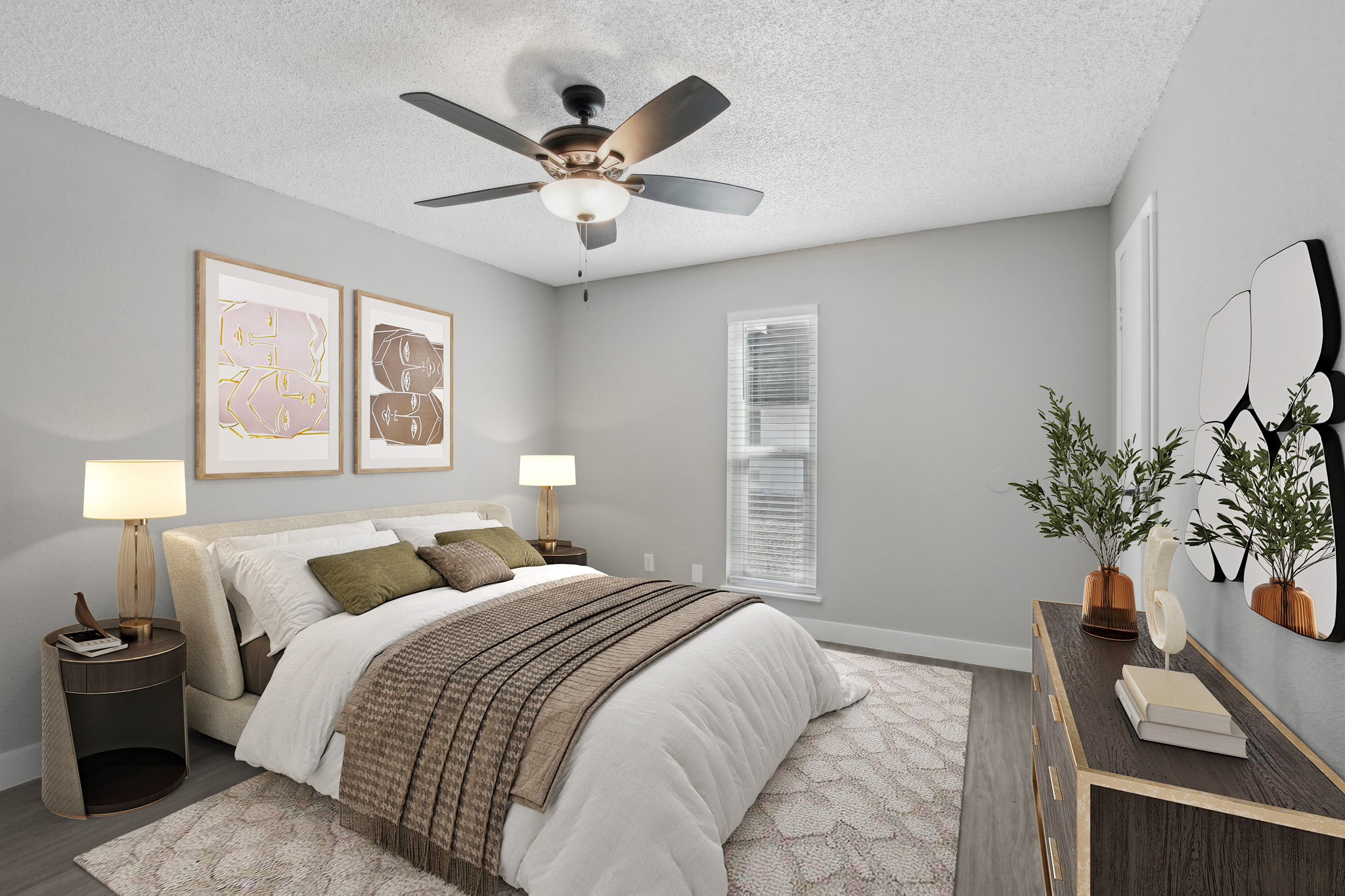 A cozy, modern bedroom featuring a queen-sized bed with neutral bedding and accent pillows. There are two framed art pieces on the wall above the bed, a nightstand with a lamp, and a dresser against the opposite wall. A ceiling fan and a window add to the bright, inviting atmosphere.
