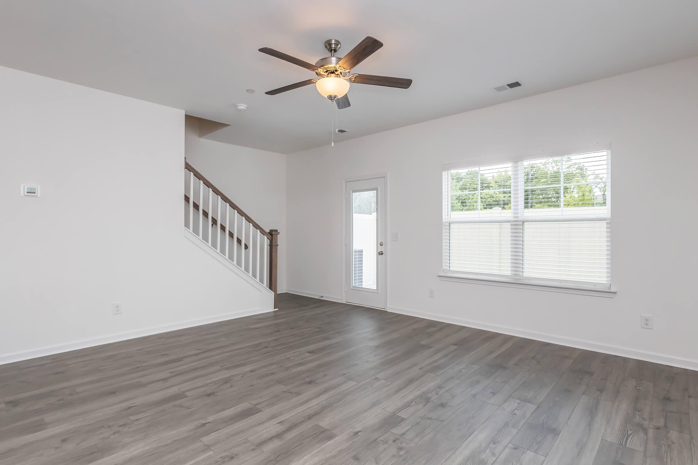 A spacious, empty living room with light-colored walls and hardwood floors. A ceiling fan hangs from the ceiling. There is a staircase in the background leading to the upper level. A door with a window beside it allows natural light, and a small window provides additional illumination.