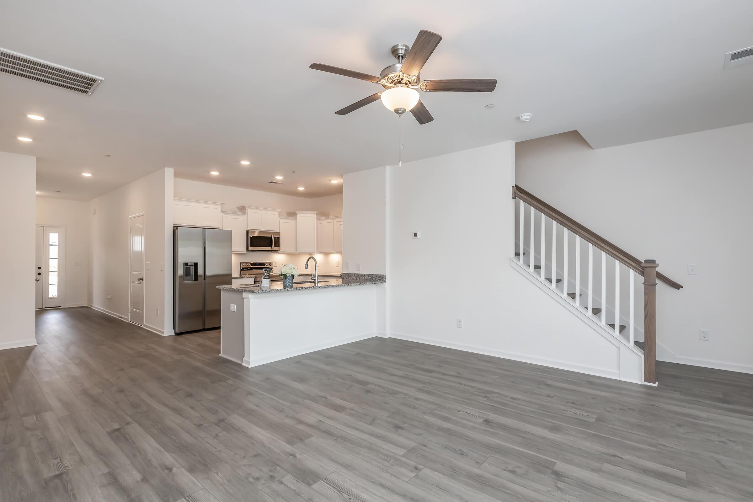 Spacious living area featuring a ceiling fan, light-colored walls, and wood-style flooring. An open kitchen with modern appliances and a large countertop is visible. A staircase leads to an upper level. Natural light comes from multiple windows, creating a bright and inviting atmosphere.