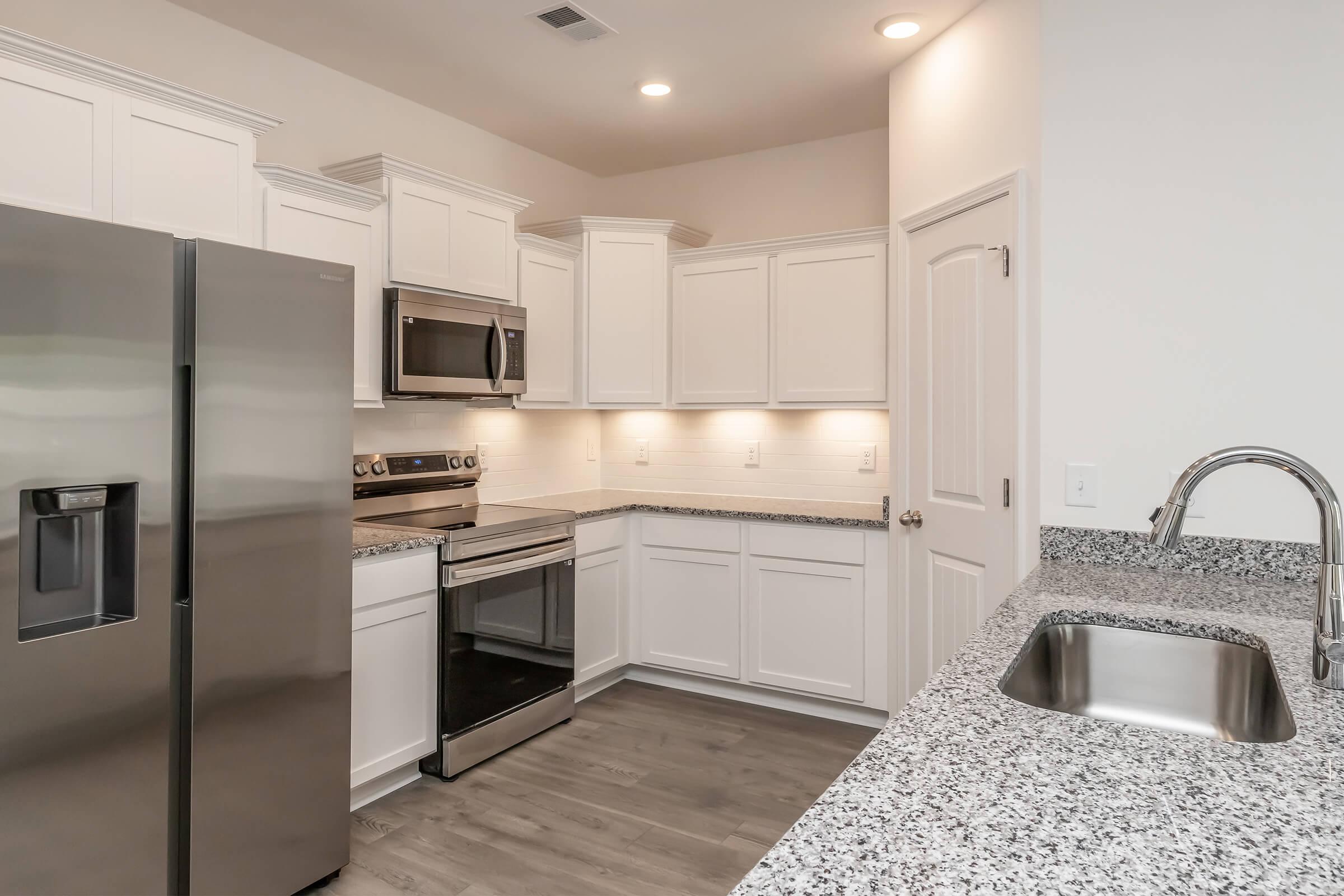 Modern kitchen featuring stainless steel appliances, including a refrigerator, oven, and microwave. The space has white cabinetry with granite countertops and a sleek sink. Soft lighting illuminates the area, highlighting the clean, minimalist design and wood-style flooring.
