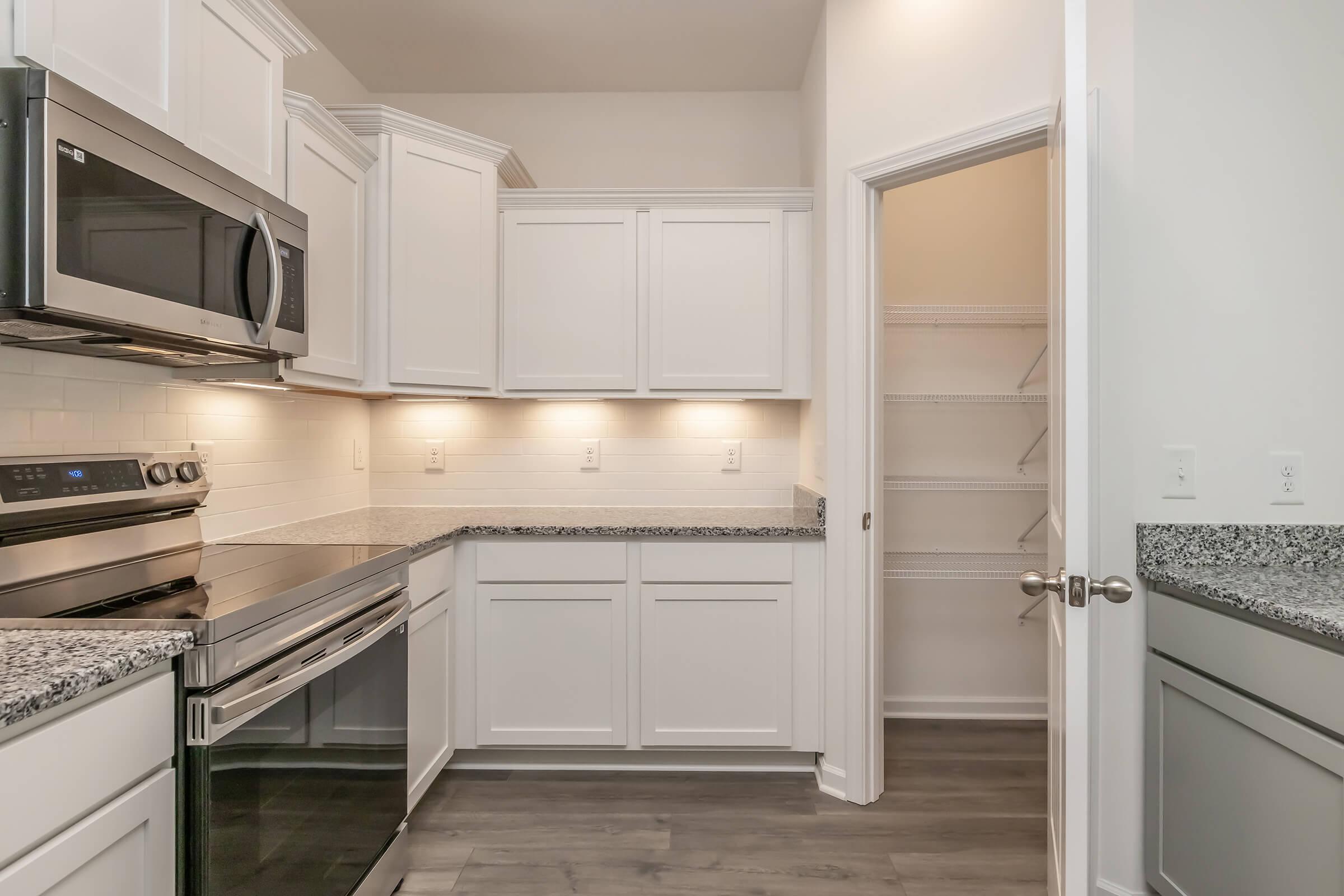 Modern kitchen featuring white cabinetry, stainless steel appliances, and granite countertops. Soft lighting illuminates the space, with a pantry visible through an open door. The flooring is a sleek dark wood, providing a stylish contrast to the bright kitchen elements.