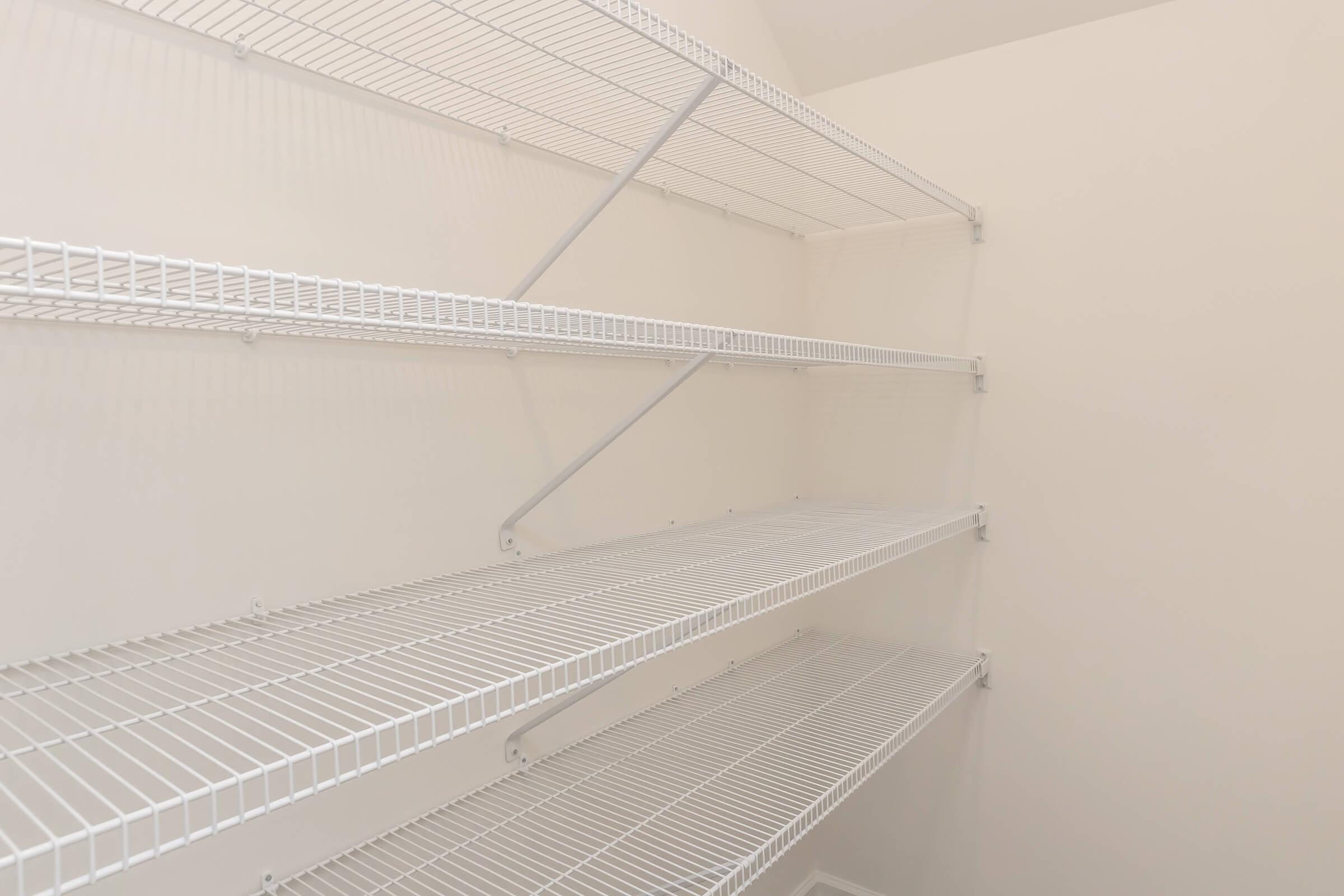 Empty white wire shelving unit attached to a light-colored wall. The shelves are designed for storage and have a minimalistic look, with multiple levels for organizing items. The background is plain and bright, emphasizing the simplicity of the shelving.