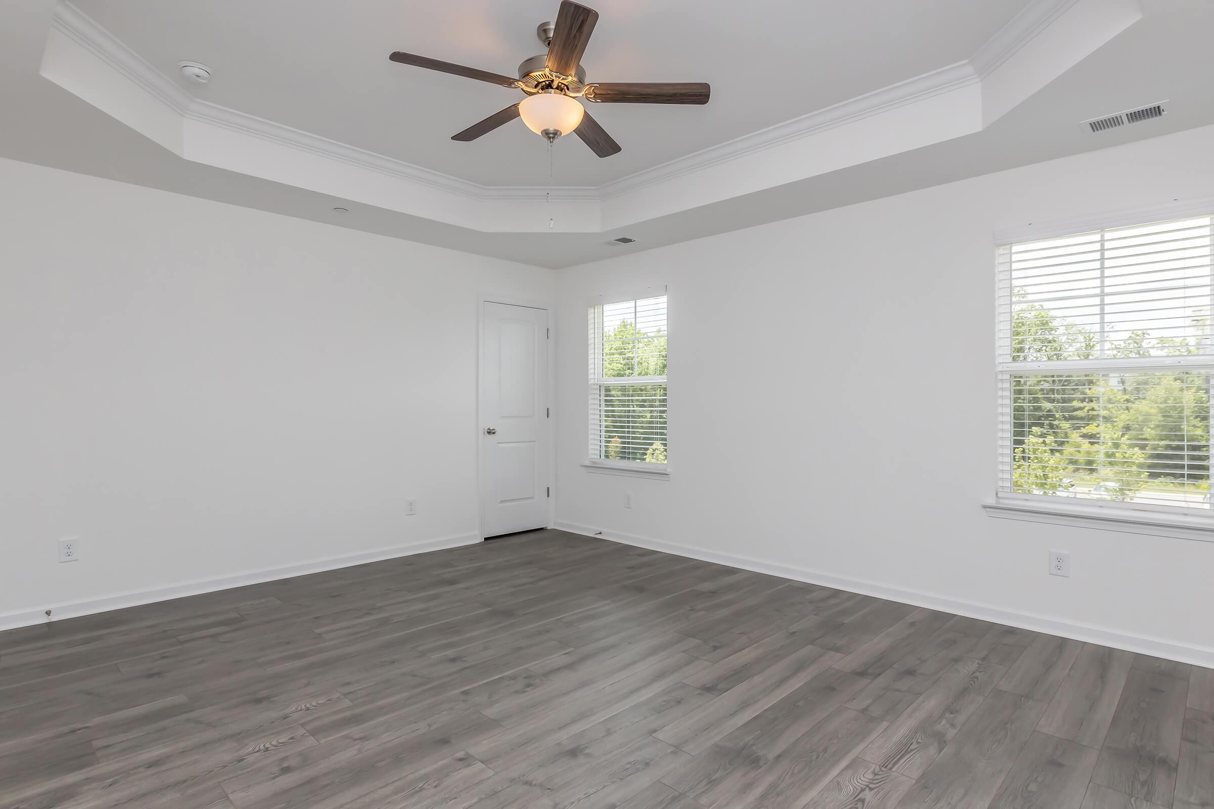 A well-lit room featuring a ceiling fan, light-colored walls, and large windows with white blinds. The floor is a modern gray laminate, and there is a single door leading to another area, creating a spacious and airy atmosphere.