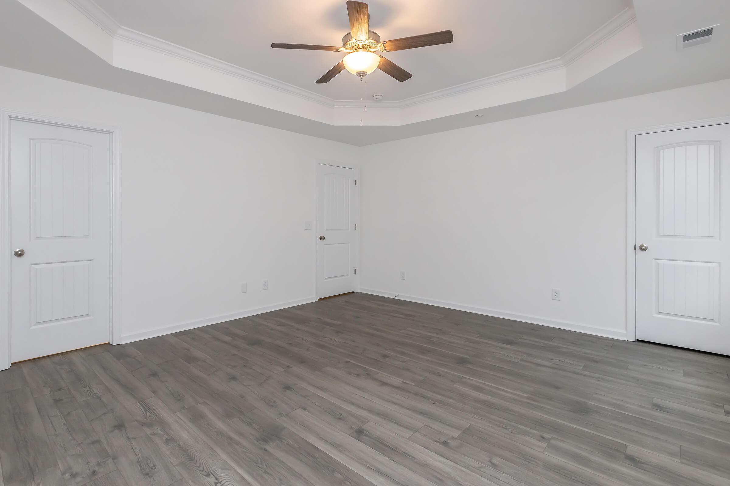 A spacious room with light gray walls and a ceiling fan. The floor is made of light-colored laminate wood. Two closed doors are visible, one on the left and one on the right. The room features a coffered ceiling, adding a decorative element to the simple design. There are no visible furnishings.