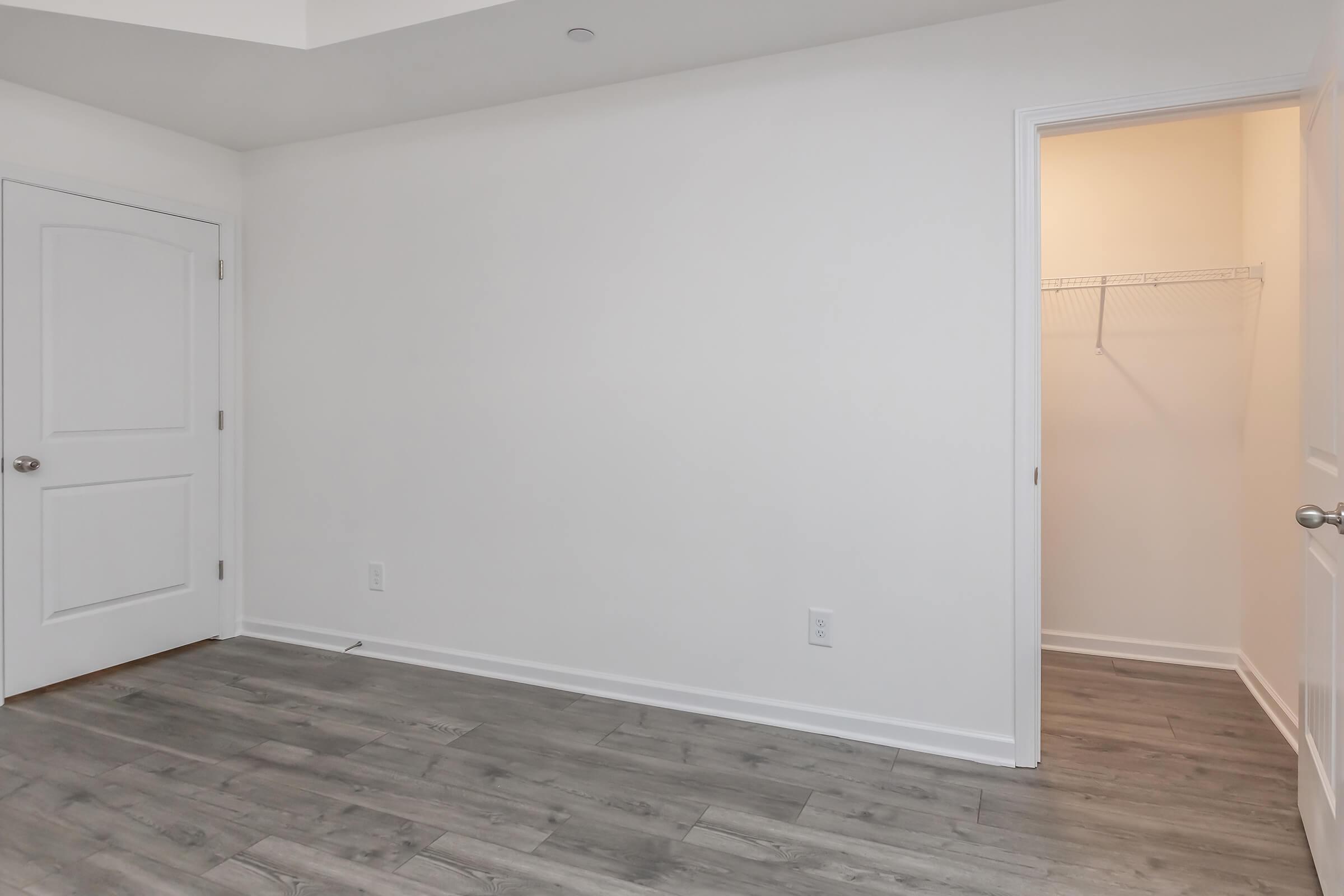 A spacious, empty room featuring light gray flooring and white walls. One side has a door leading to a closet with a wire shelf, while the other side is open with a doorway. The ceiling is bright, and the overall space looks modern and minimalistic.