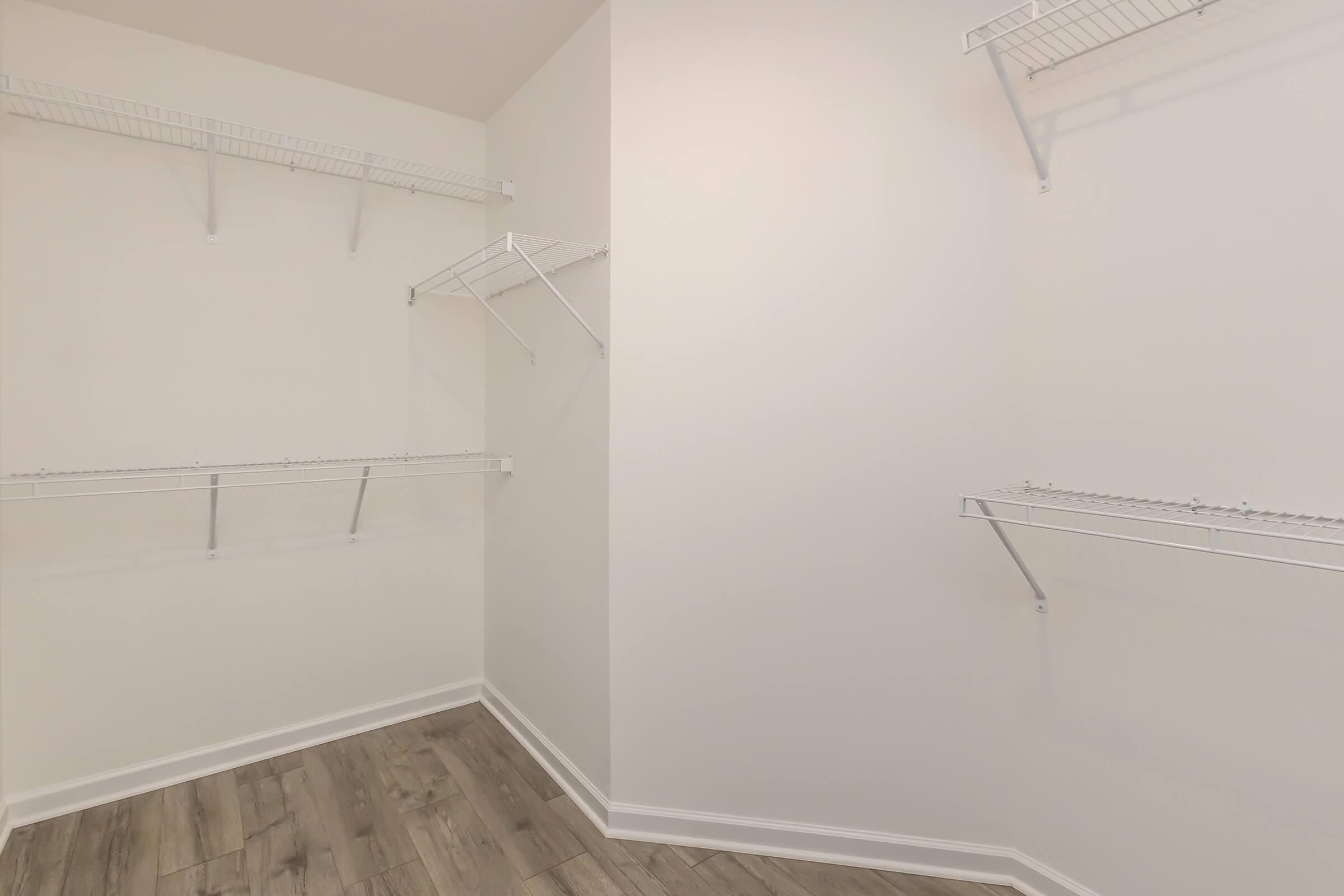 Empty closet with white wire shelving mounted on light-colored walls. The floor has wooden planks, and the space is well-lit, creating a clean and organized appearance. The shelving is positioned to maximize storage options in the corner of the room.