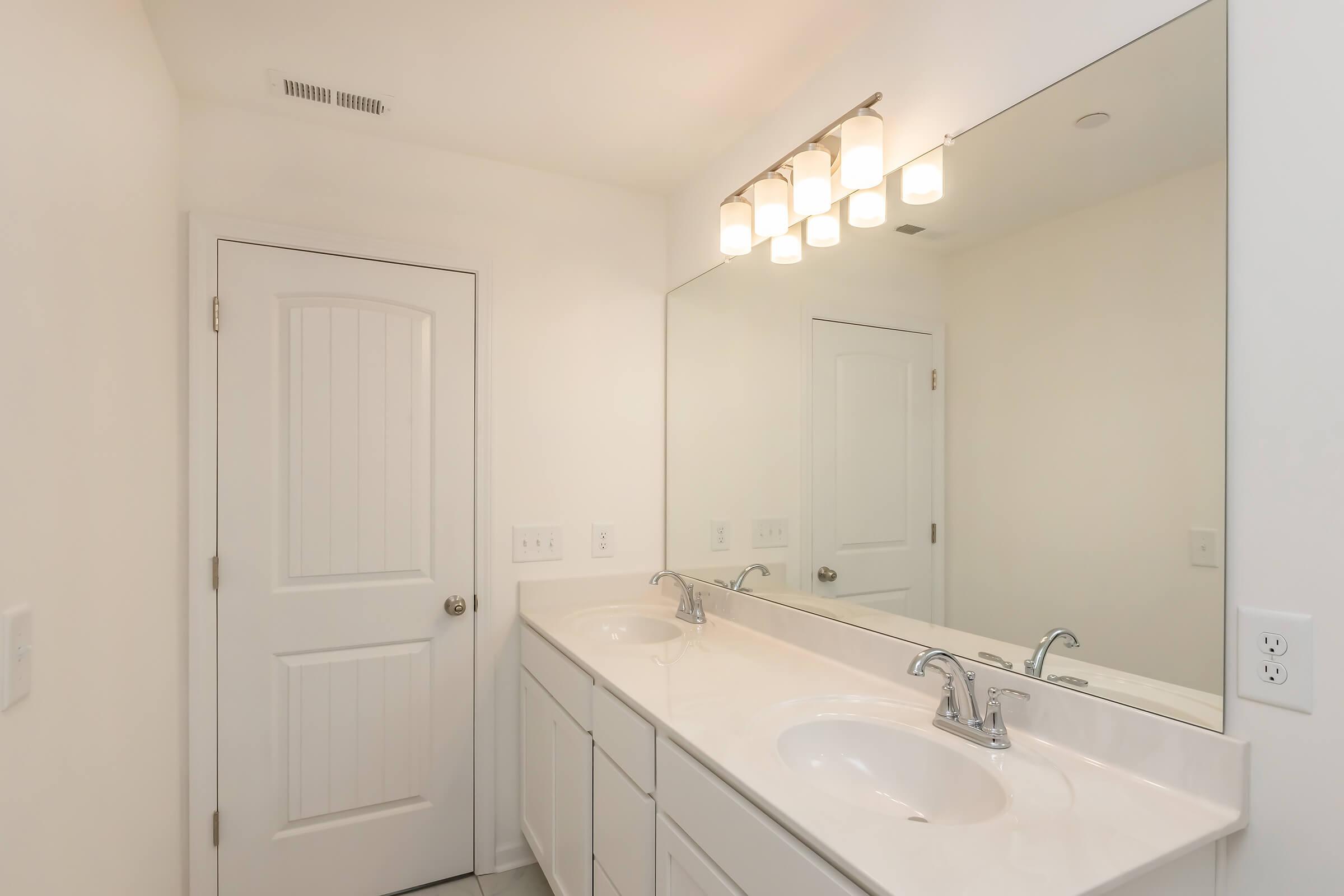 Bright and modern bathroom featuring double sinks with silver faucets, a large mirror above the sinks, and a white door. The walls and cabinetry are painted in a light color, creating a clean and spacious look. Soft overhead lighting enhances the ambiance, making the space feel welcoming and well-lit.