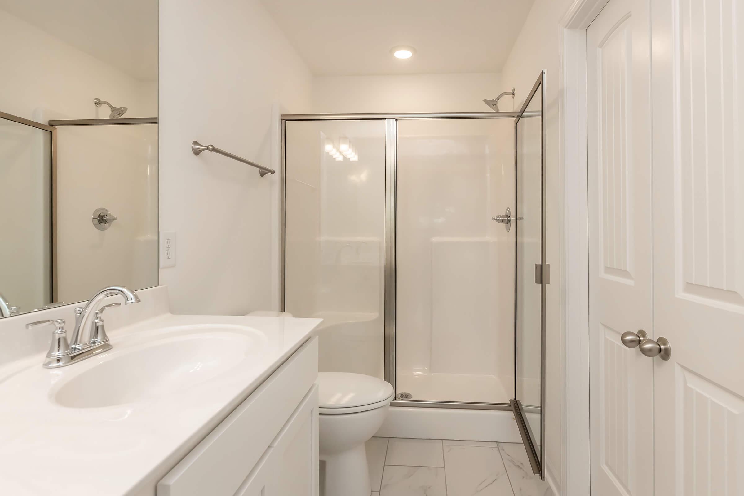 A modern bathroom featuring a glass-enclosed shower, a white sink with chrome faucet, and a toilet. The walls are painted in a light color, and the flooring consists of large white tiles. A large mirror above the sink reflects the clean and minimalistic design.