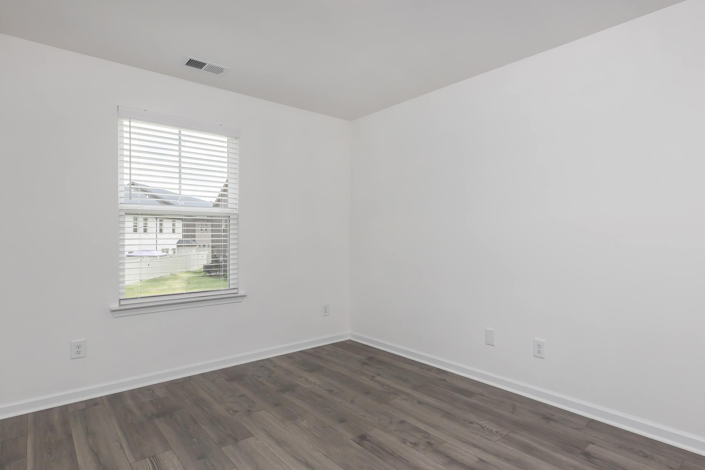 A clean, empty room featuring light-colored walls and a large window with white blinds. The floor is made of light wood laminate. Natural light enters through the window, illuminating the spacious, minimalistic interior.