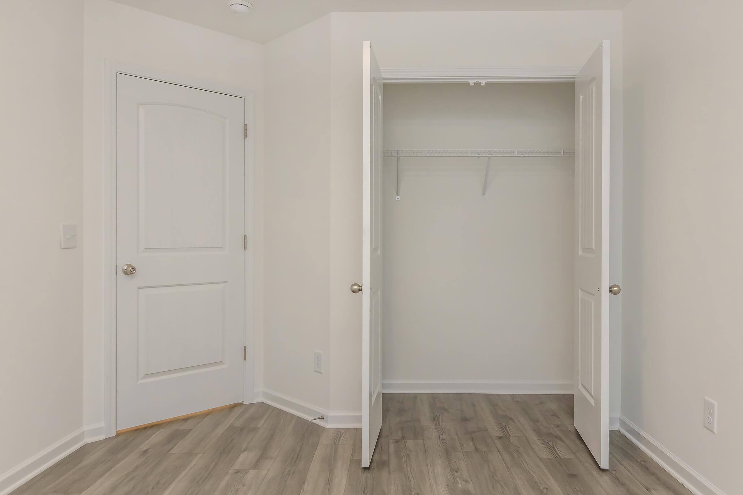 A clean, neutral-colored room featuring a partially open closet with a hanging rod, light wooden flooring, and a closed door on the left. The walls are painted white, creating a bright and airy atmosphere.
