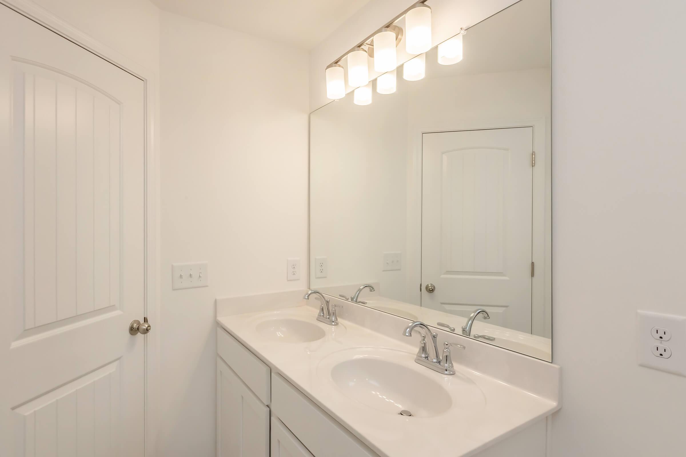 A bright bathroom featuring a double sink vanity with two sinks and modern faucets. Above the sinks, there is a row of five light fixtures. The walls are plain white, and a door is visible in the background, adding to the clean and spacious feel of the room.