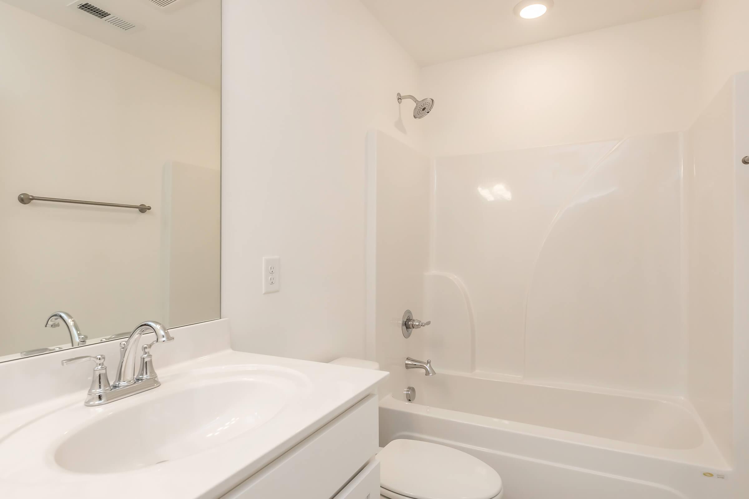 A clean, modern bathroom featuring a white bathtub and shower combination, a white sink with a chrome faucet, and a large mirror. The walls are painted a light color, and there are simple fixtures, creating a bright and spacious atmosphere.