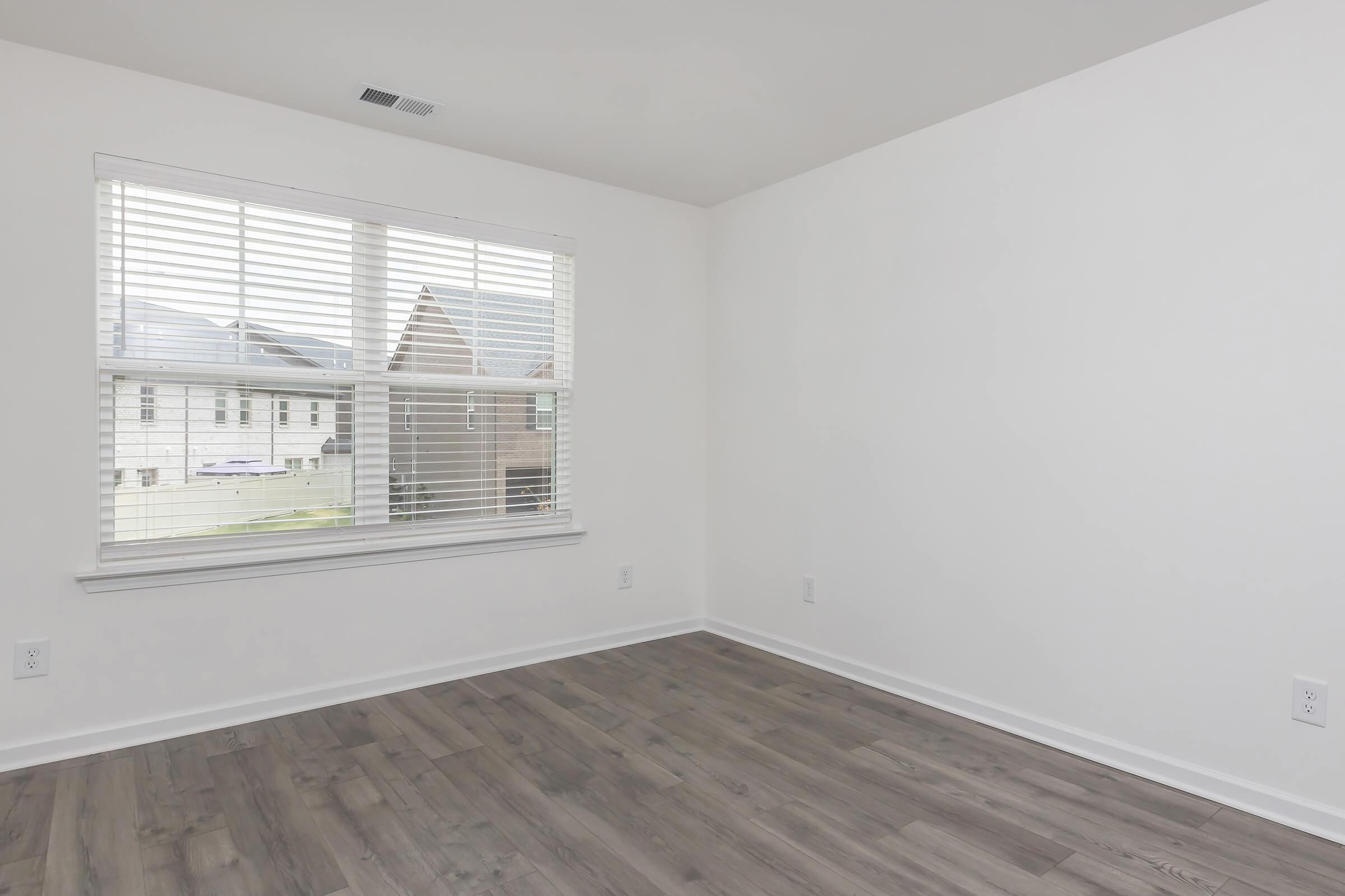 A bright, empty room with light-colored walls and wooden flooring. A large window with white blinds lets in natural light, revealing a view outside of adjacent houses. The space is minimalistic and ready for furnishing.