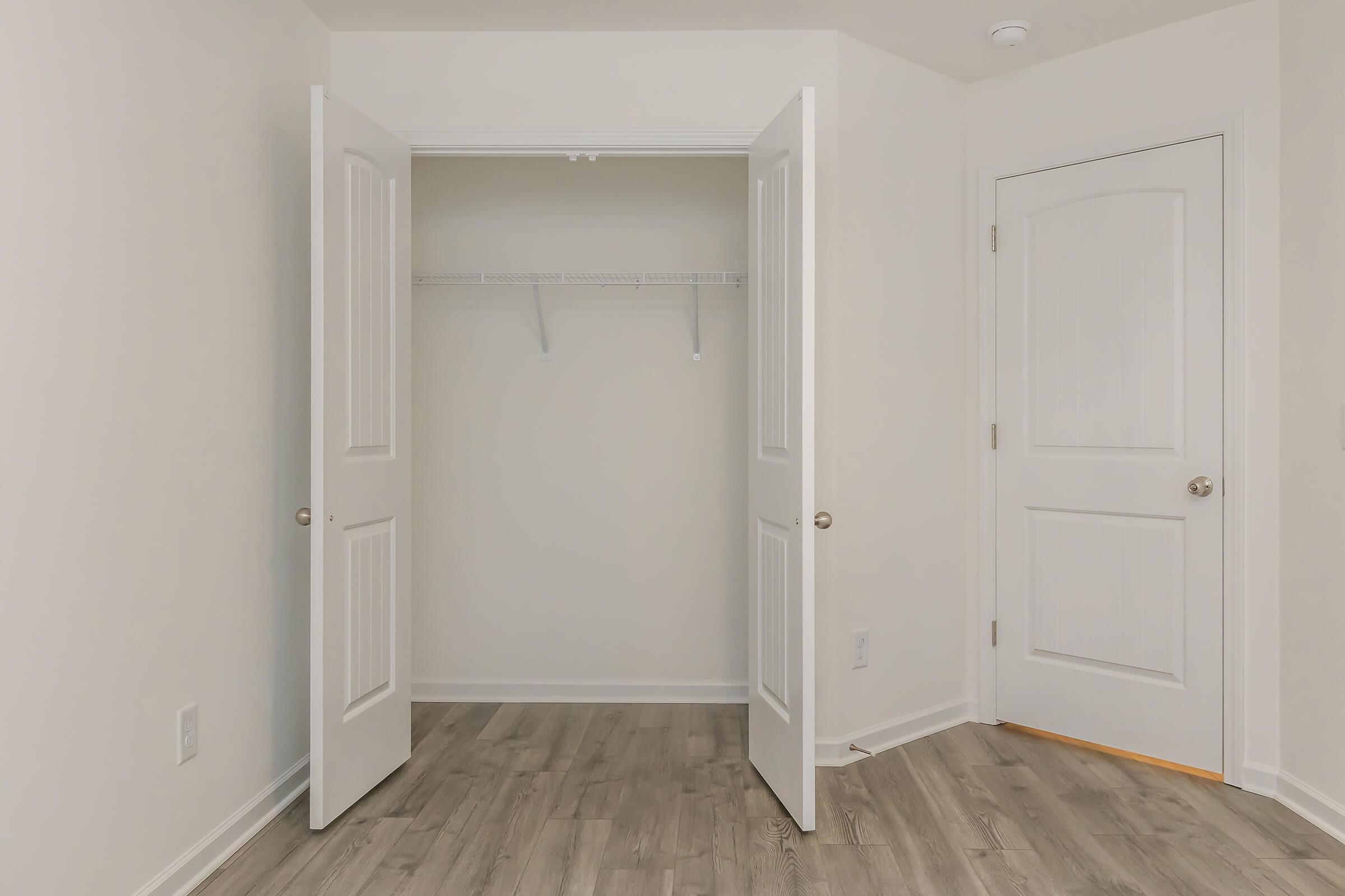Interior of a room featuring two open doors leading to a closet. The closet has a wire shelf along the back wall. The walls are painted a light color, and the floor has a light brown wood-like finish. There is a ceiling light visible in the corner.