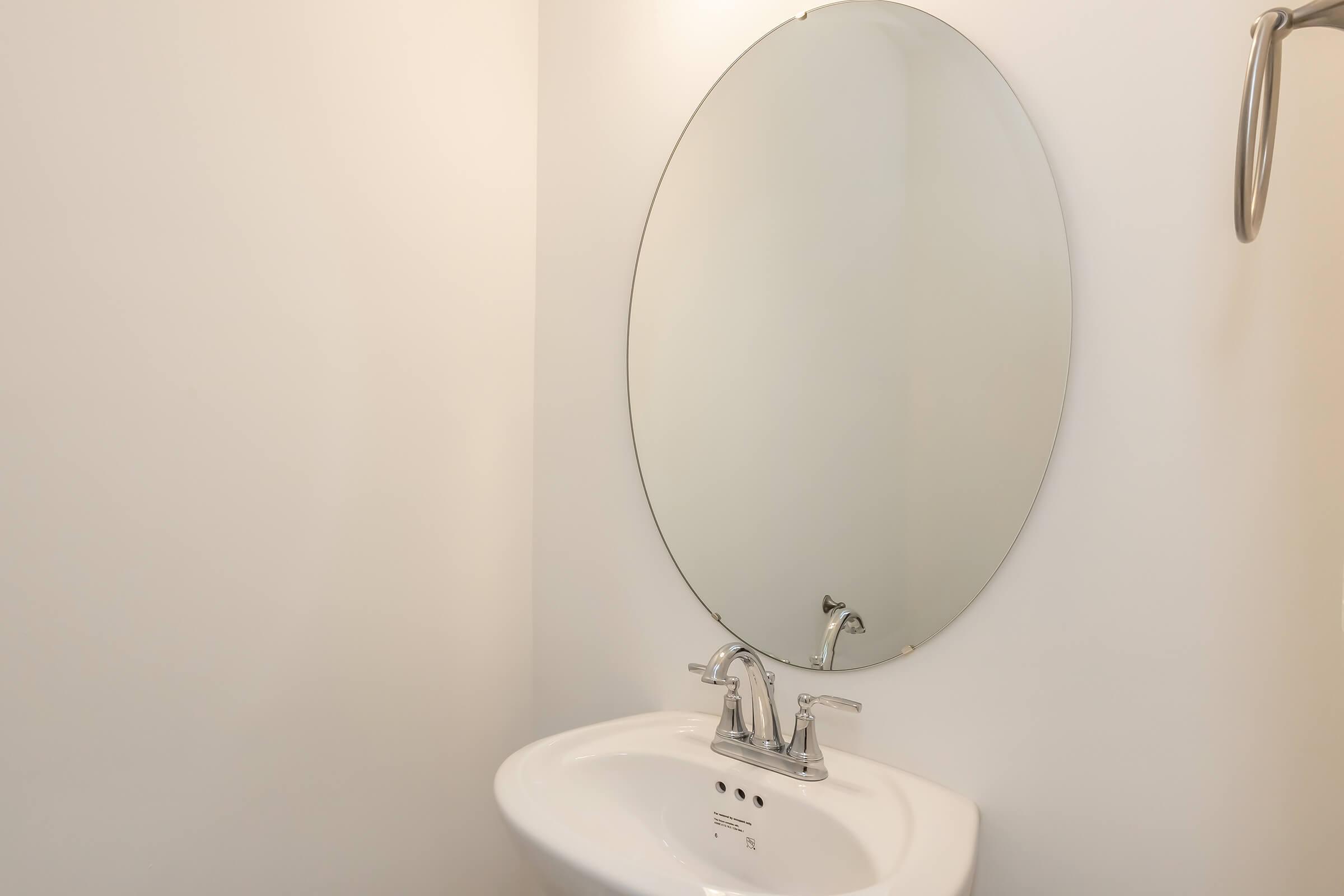 A clean and minimalist bathroom setting featuring a modern oval mirror above a sleek white sink. The surrounding walls are painted in a light color, enhancing the airy feel of the space. The faucet is contemporary, and a towel holder is mounted on the wall nearby.