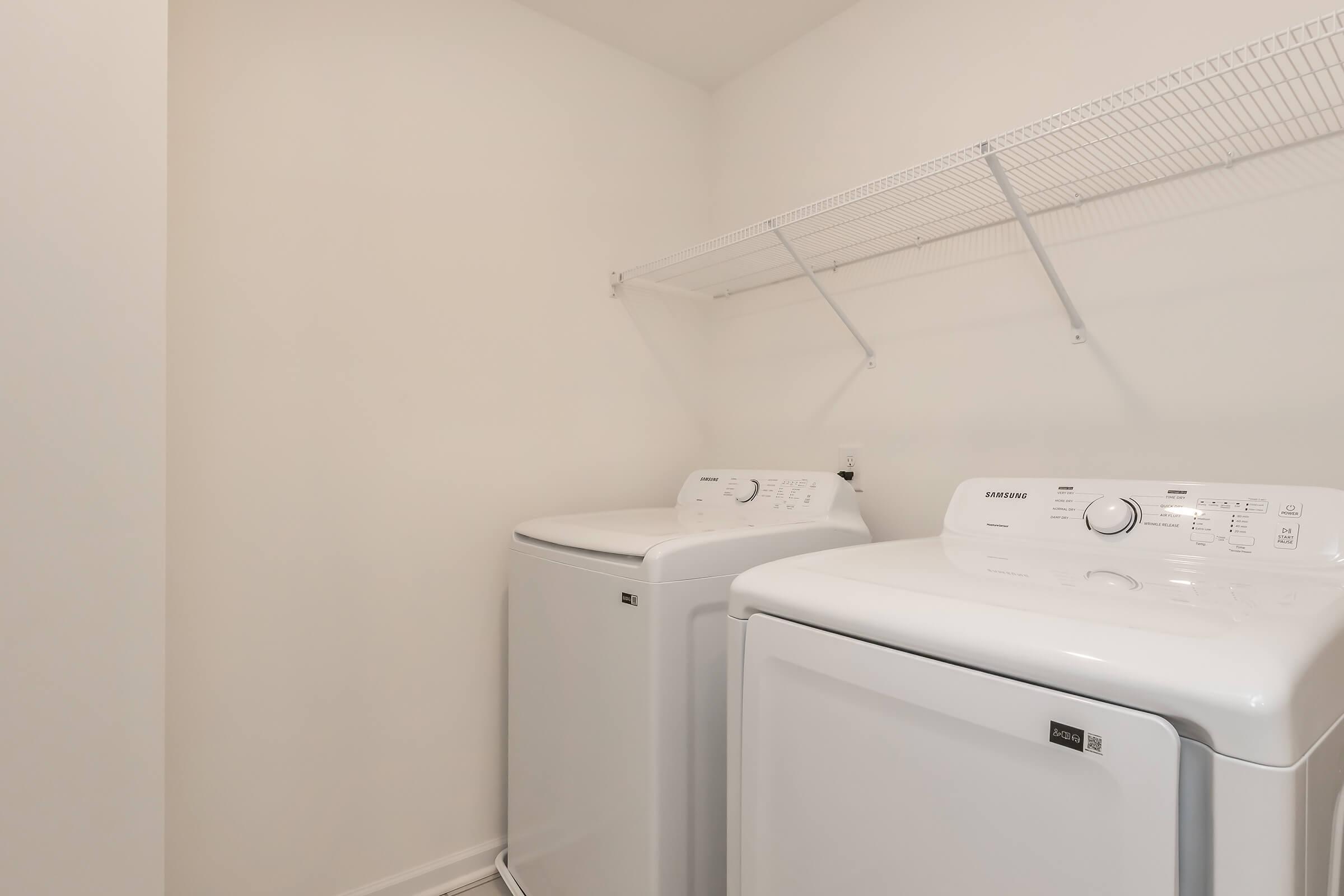 A clean laundry room featuring two white washing machines side by side. Above them is a metal shelving unit for storage, with plain white walls providing a bright and simple atmosphere. The space is organized and uncluttered, suitable for laundry tasks.