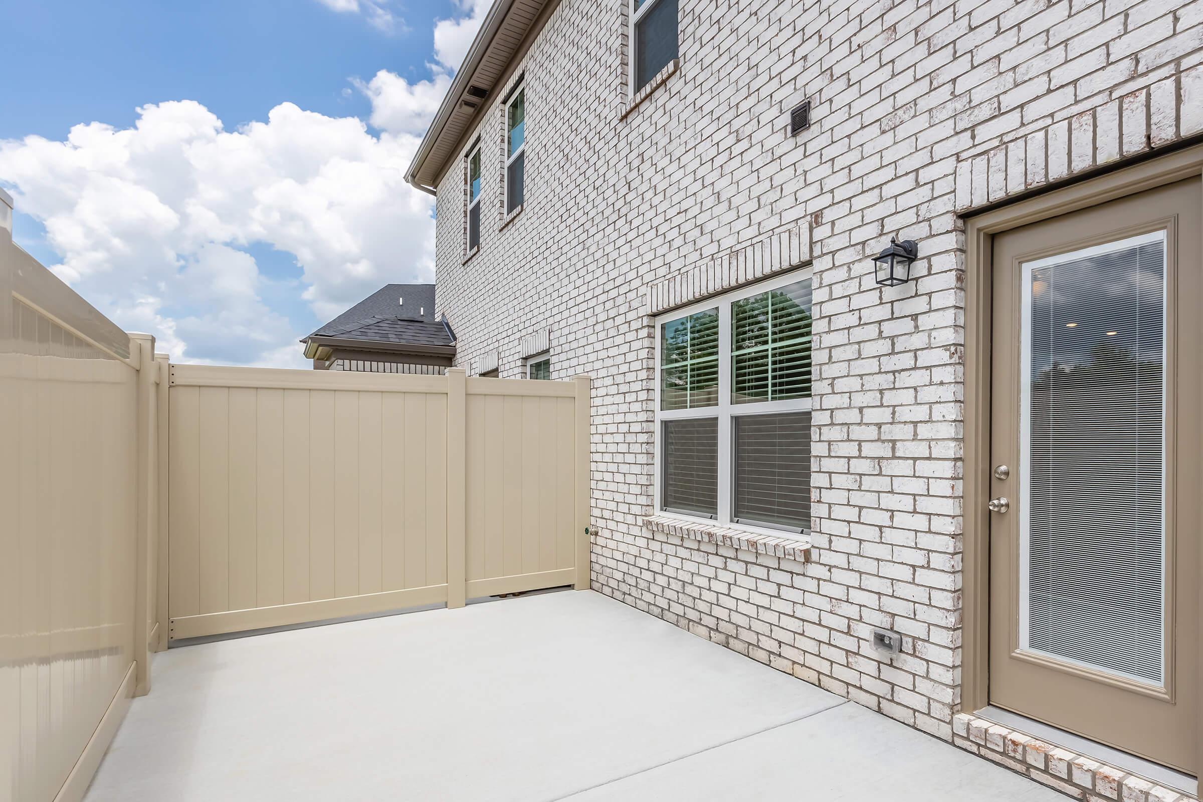 A modern exterior view of a house featuring a white brick wall, a beige privacy fence, and a concrete patio area. The house has large windows and a door leading to the outside. The sky is bright with scattered clouds, enhancing the overall inviting atmosphere of the outdoor space.