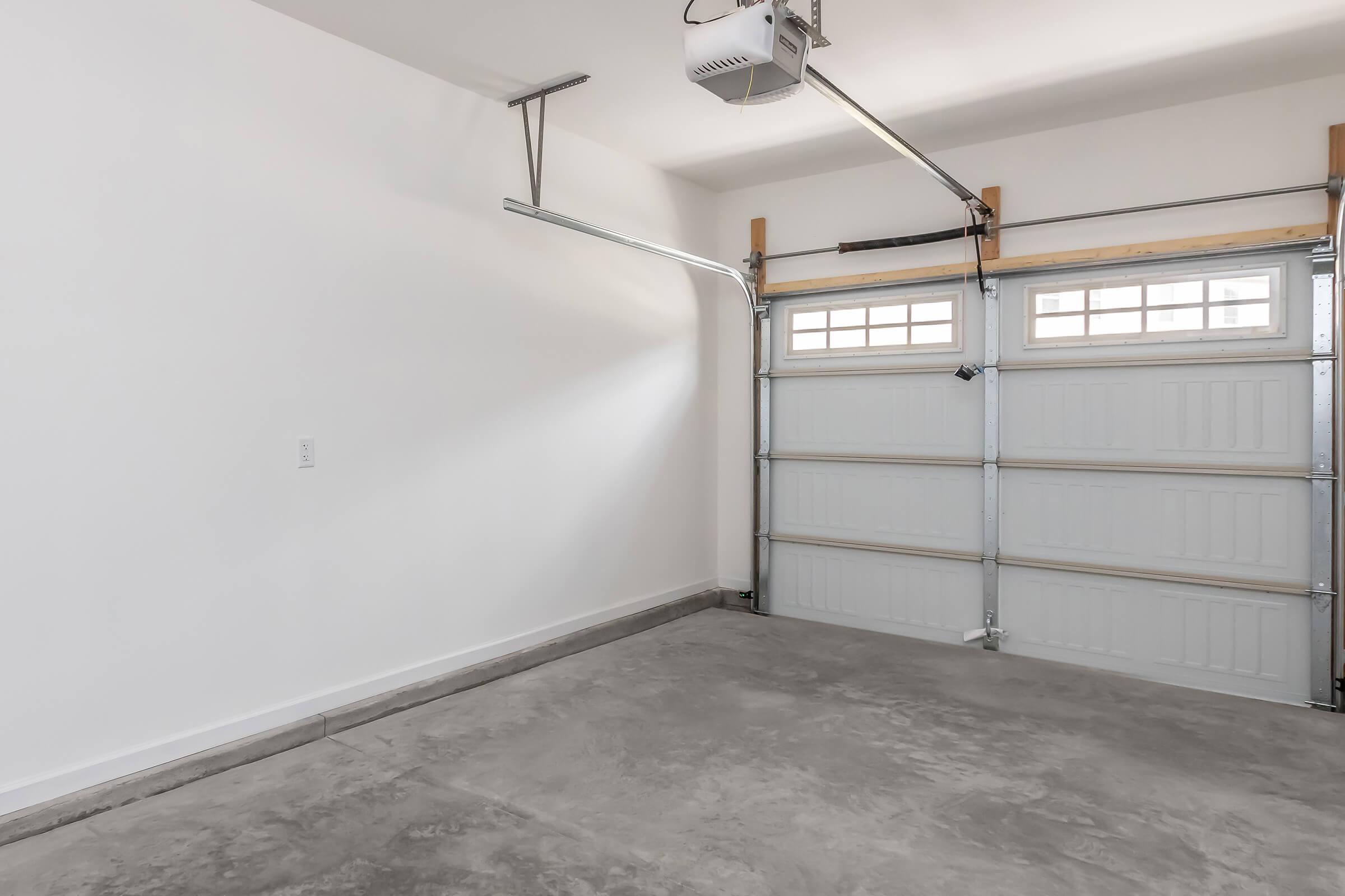 A clean, empty garage with a polished concrete floor and a large garage door. The walls are painted white, and the garage door is partially closed with windows at the top. There is an overhead garage door opener mounted on the ceiling. The space is well-lit and uncluttered.