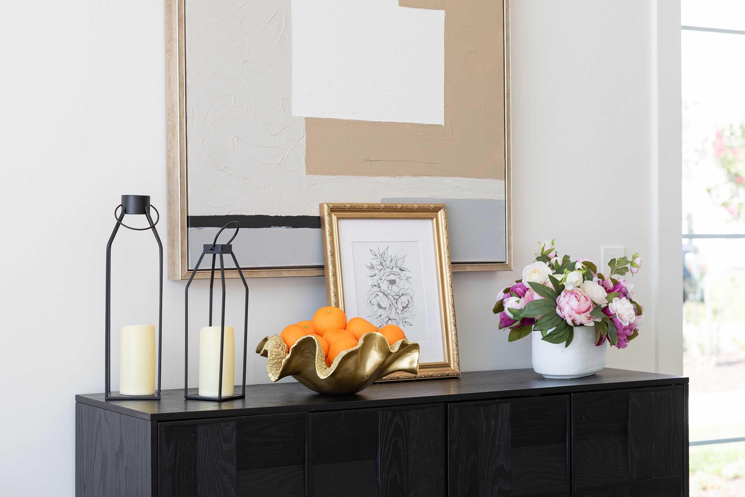 A stylish interior arrangement featuring a dark wooden console table topped with a gold decorative bowl of oranges, a white vase filled with pink and white flowers, two black lanterns with candles, and a framed artwork hanging on the wall above.