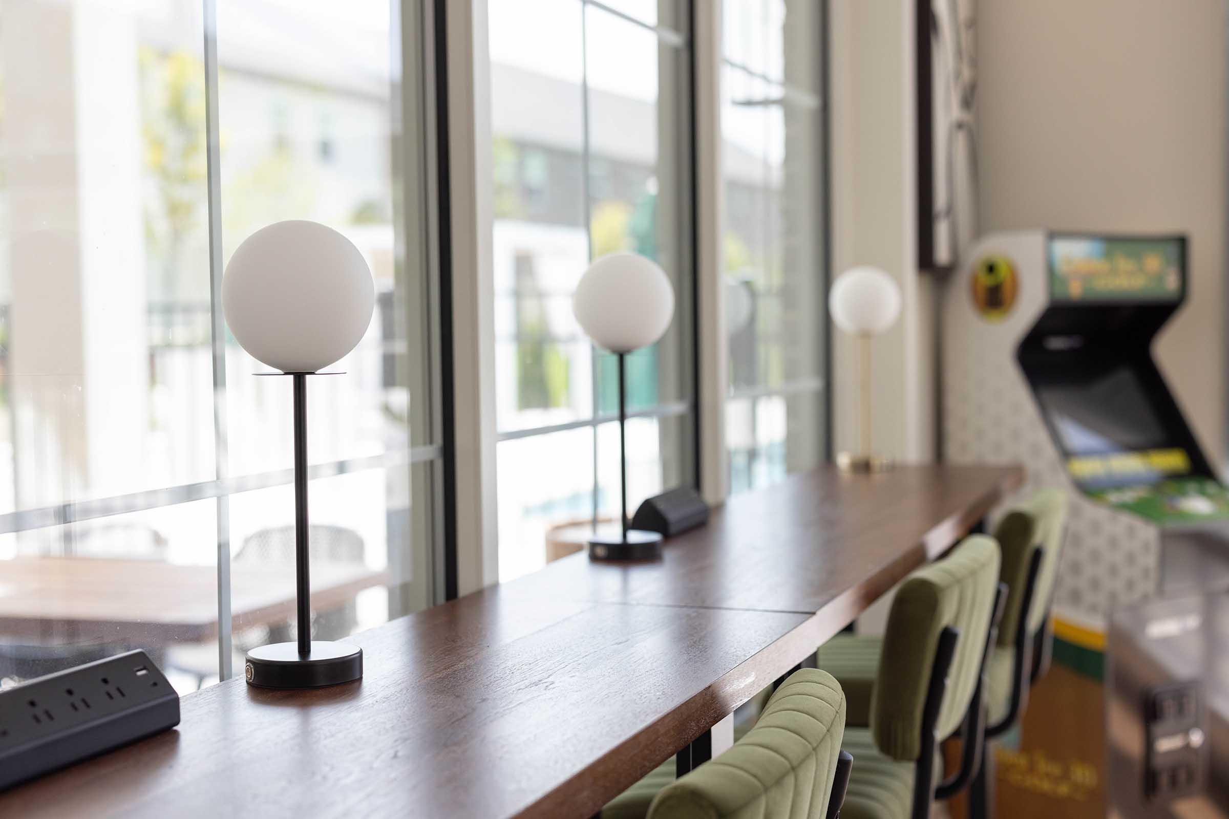 A well-lit interior space featuring a wooden table with three modern table lamps and green upholstered chairs. In the background, there is an arcade machine, suggesting a recreational area. Large windows allow natural light to fill the room, enhancing the inviting atmosphere.
