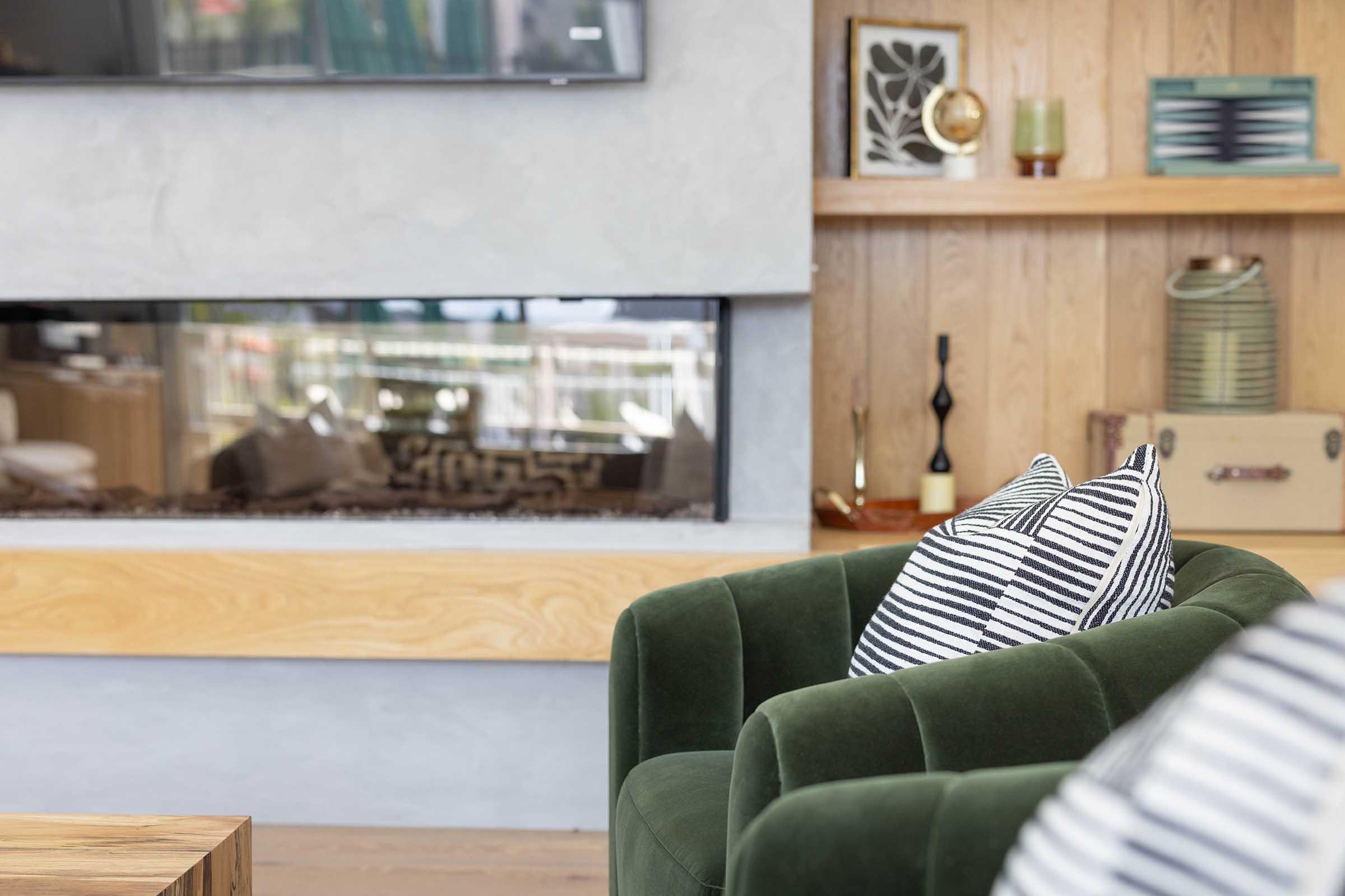 A cozy living room with a green velvet armchair, featuring a striped pillow. In the background, there's a modern fireplace built into a wooden wall, adorned with decorative items like a vase and framed artwork. Soft natural light fills the room, creating a warm and inviting atmosphere.