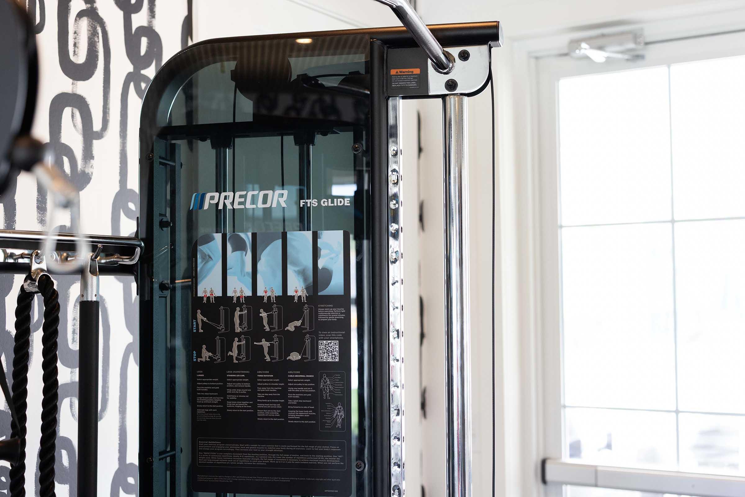 A close-up view of a Precor FTS Glide strength training machine, featuring a sleek design with transparent panels and a detailed exercise instructions placard. The background includes a bright window and a patterned wall, suggesting a modern gym environment.