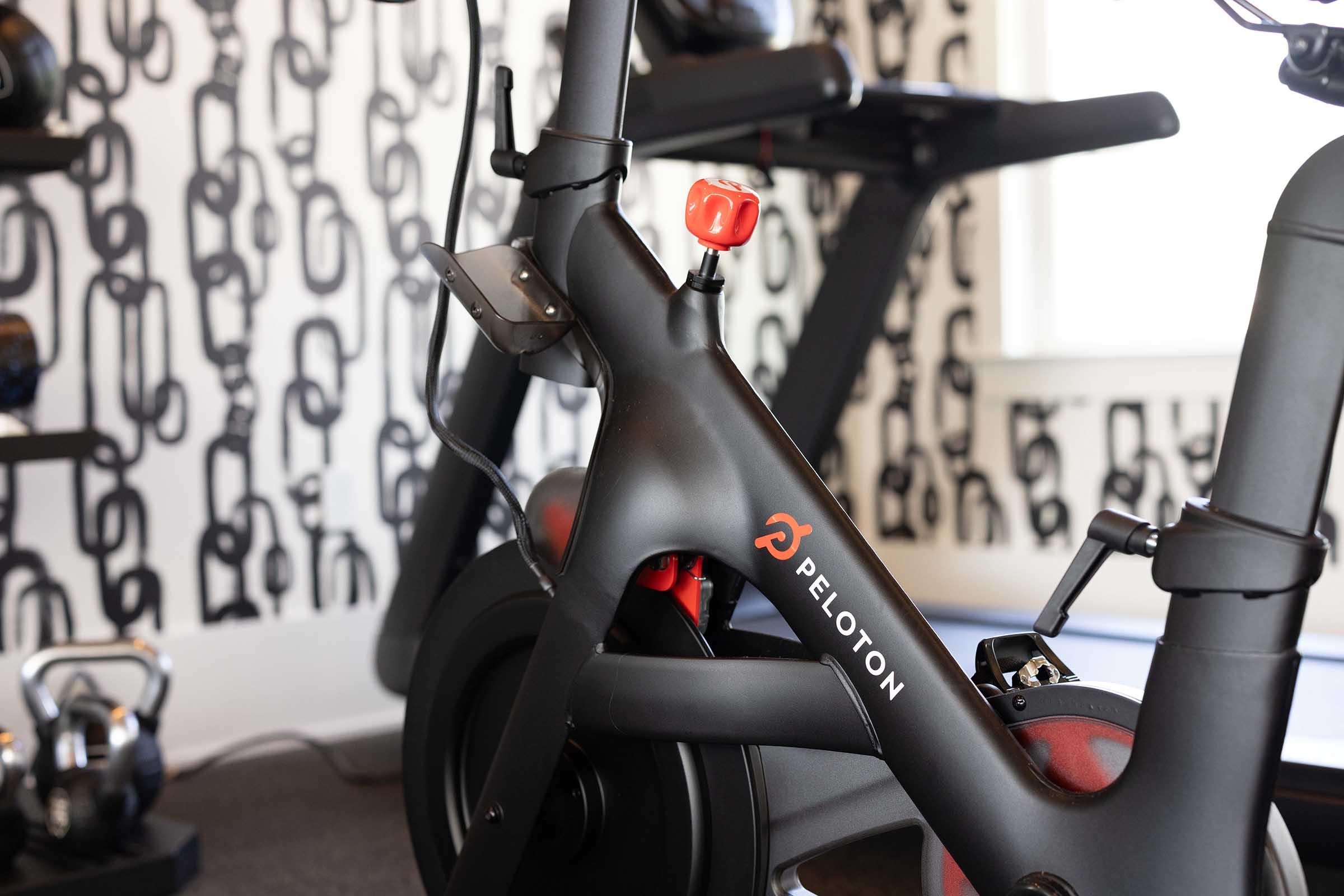 A close-up of a Peloton exercise bike, showcasing the frame and resistance knob. The background features a patterned wall with abstract designs. Another exercise machine is partially visible, indicating a fitness environment.