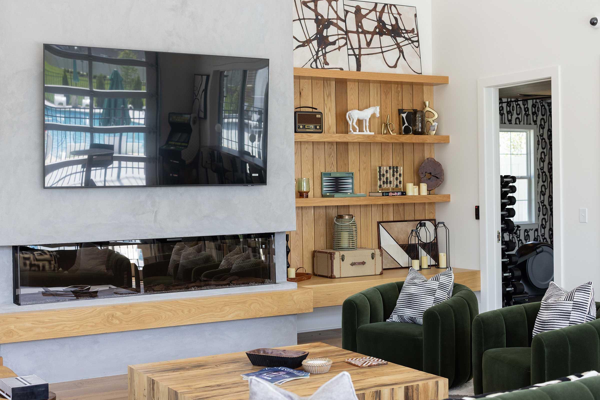 A modern living room featuring a mounted flat-screen TV above a sleek fireplace. There is a wooden shelf displaying decorative items and a white toy horse. Comfortable green armchairs are arranged around a wooden coffee table, with a view of a swimming pool visible through large windows in the background.