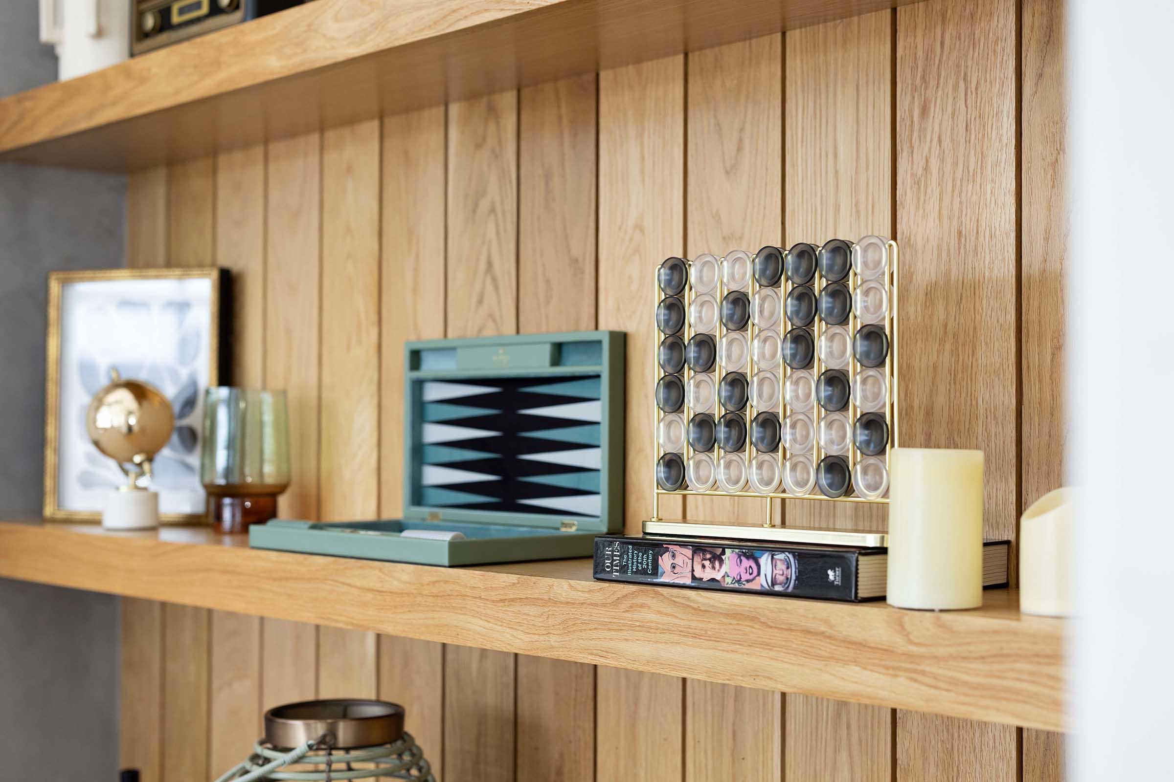 A wooden shelf displaying various decorative items including a vintage-style board game with geometric patterns, a framed photo, a small globe, a candle, and a book with a portrait on its cover. The overall aesthetic is modern and minimalistic with warm tones and different textures.