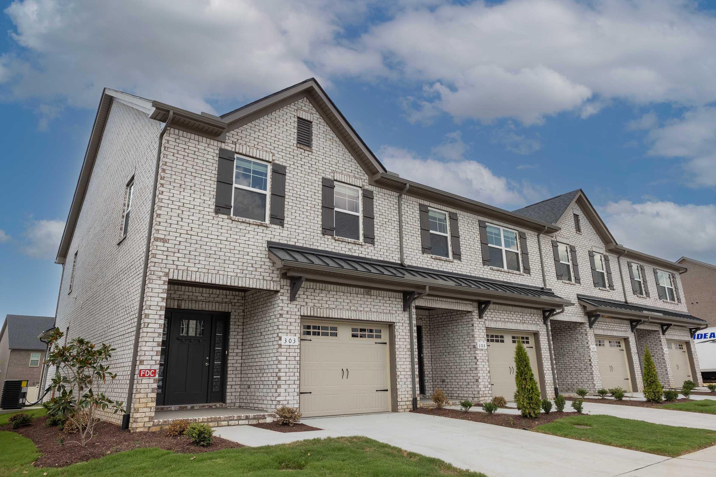 A row of modern townhouses featuring brick exteriors with gray roofs. Each unit has a garage door and well-maintained landscaping with small shrubs and grass. The sky is partly cloudy, adding a bright backdrop to the homes.