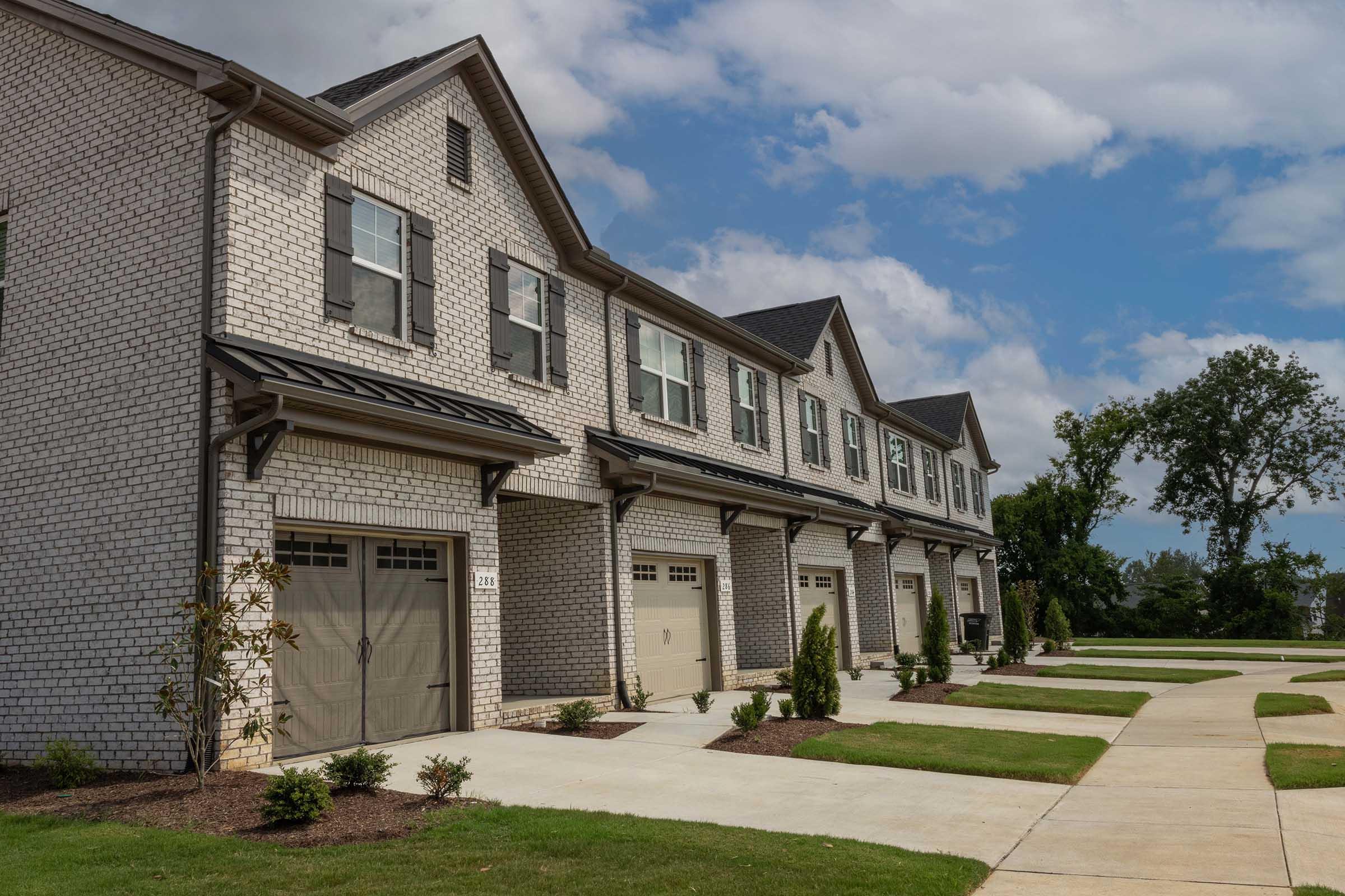 Row of brick townhouses with gray shutters and garages, set against a partly cloudy blue sky. Neatly landscaped lawn with sidewalks leading to the homes, creating a suburban residential atmosphere.