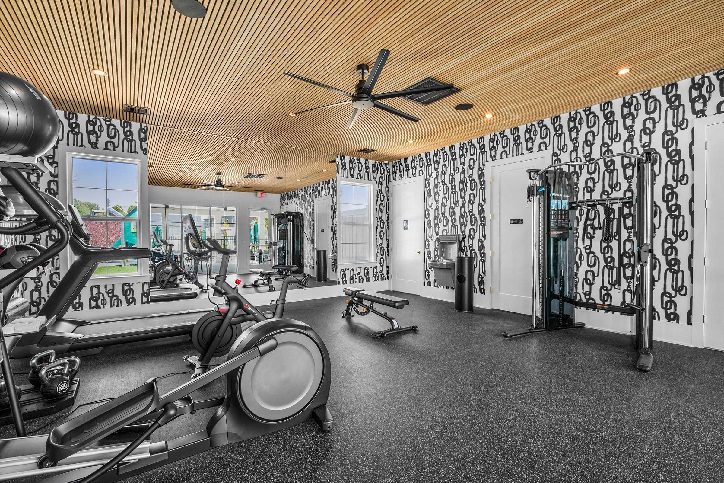 A modern fitness room featuring exercise machines like treadmills and stationary bikes, weights, and a cable machine. The room has a sleek design with a wooden ceiling, decorative wall patterns, large windows providing natural light, and a water cooler.