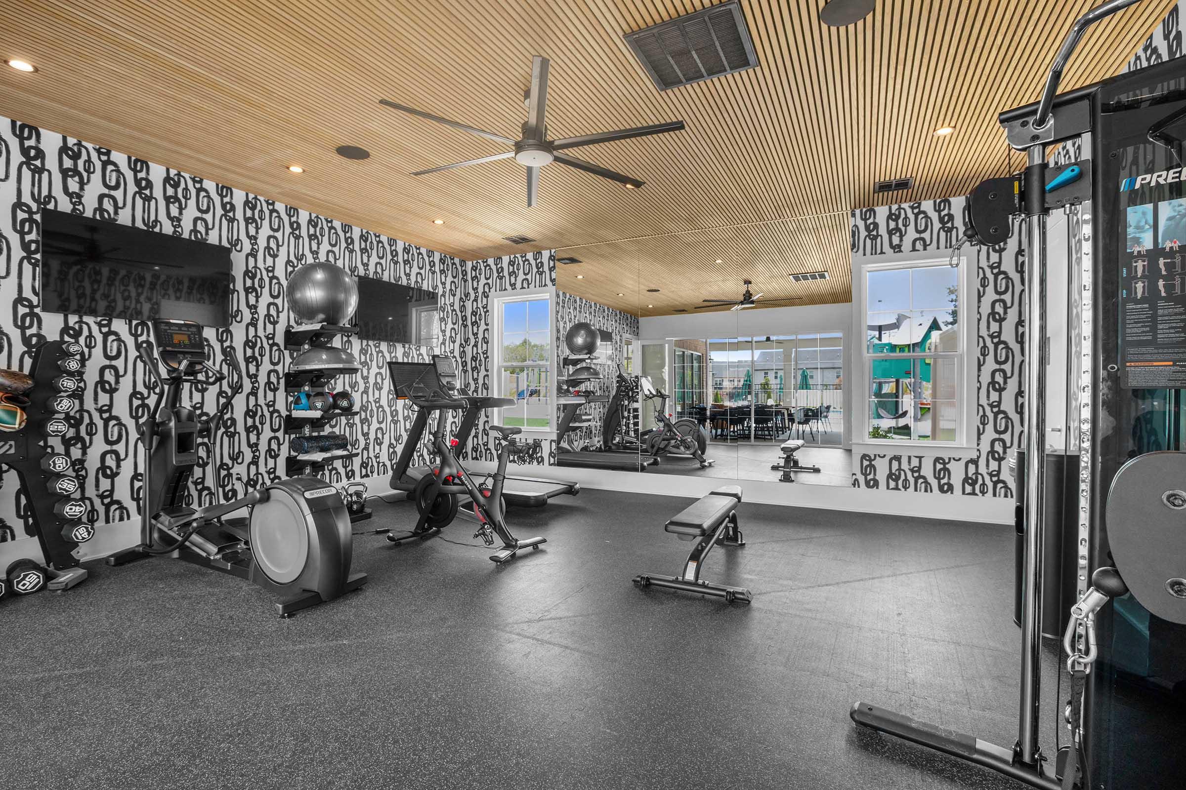 A modern gym interior featuring various fitness equipment, including a treadmill, stationary bike, weight bench, and a multi-gym station. The walls are decorated with a chain pattern, and large windows provide natural light, creating an inviting workout space.