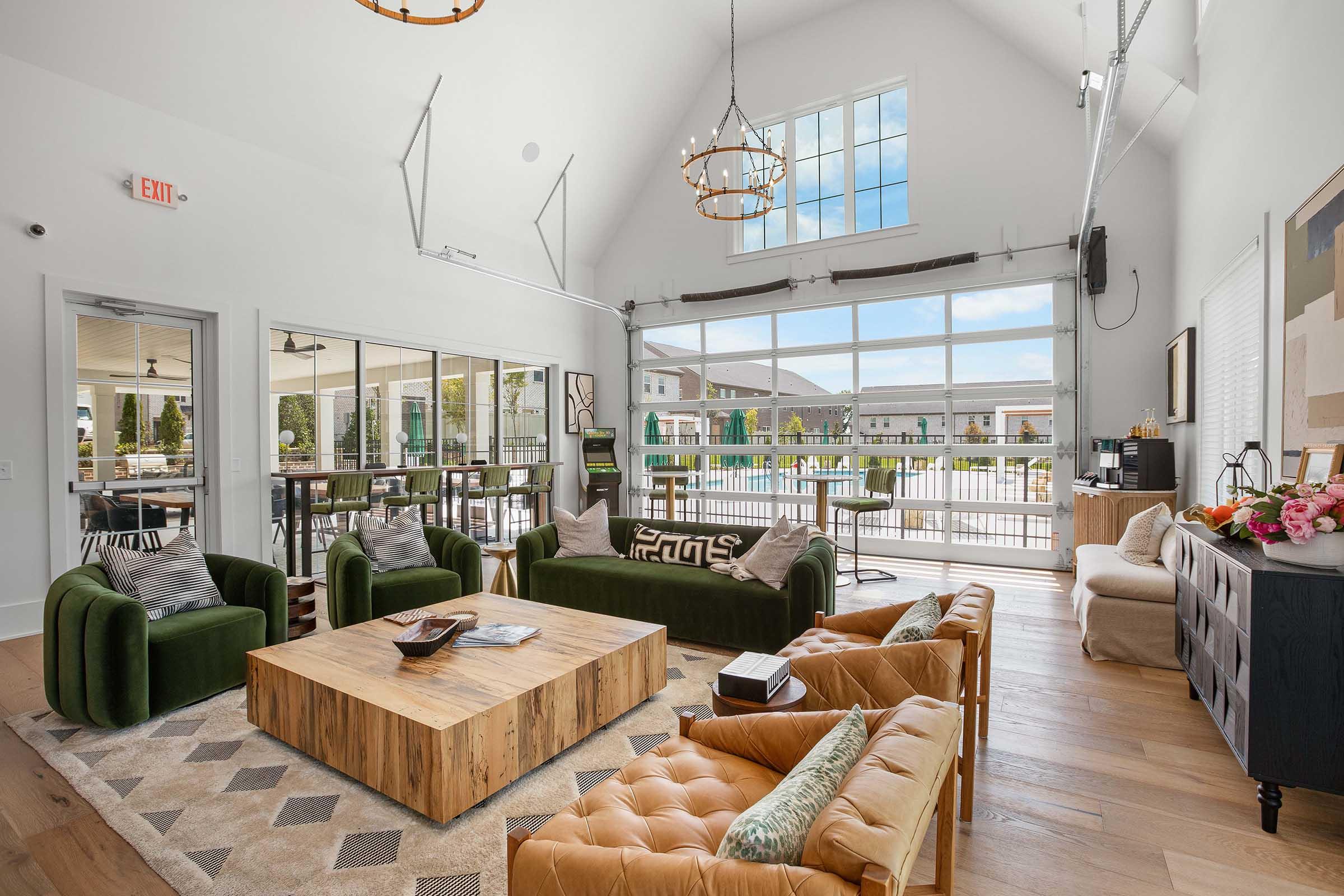A spacious and modern lounge area featuring a high ceiling, large windows, and a mix of seating options, including green sofas, a wooden coffee table, and a tan leather couch. The space is brightly lit and opens to an outdoor area with a pool view, decorated with plants and stylish lighting fixtures.