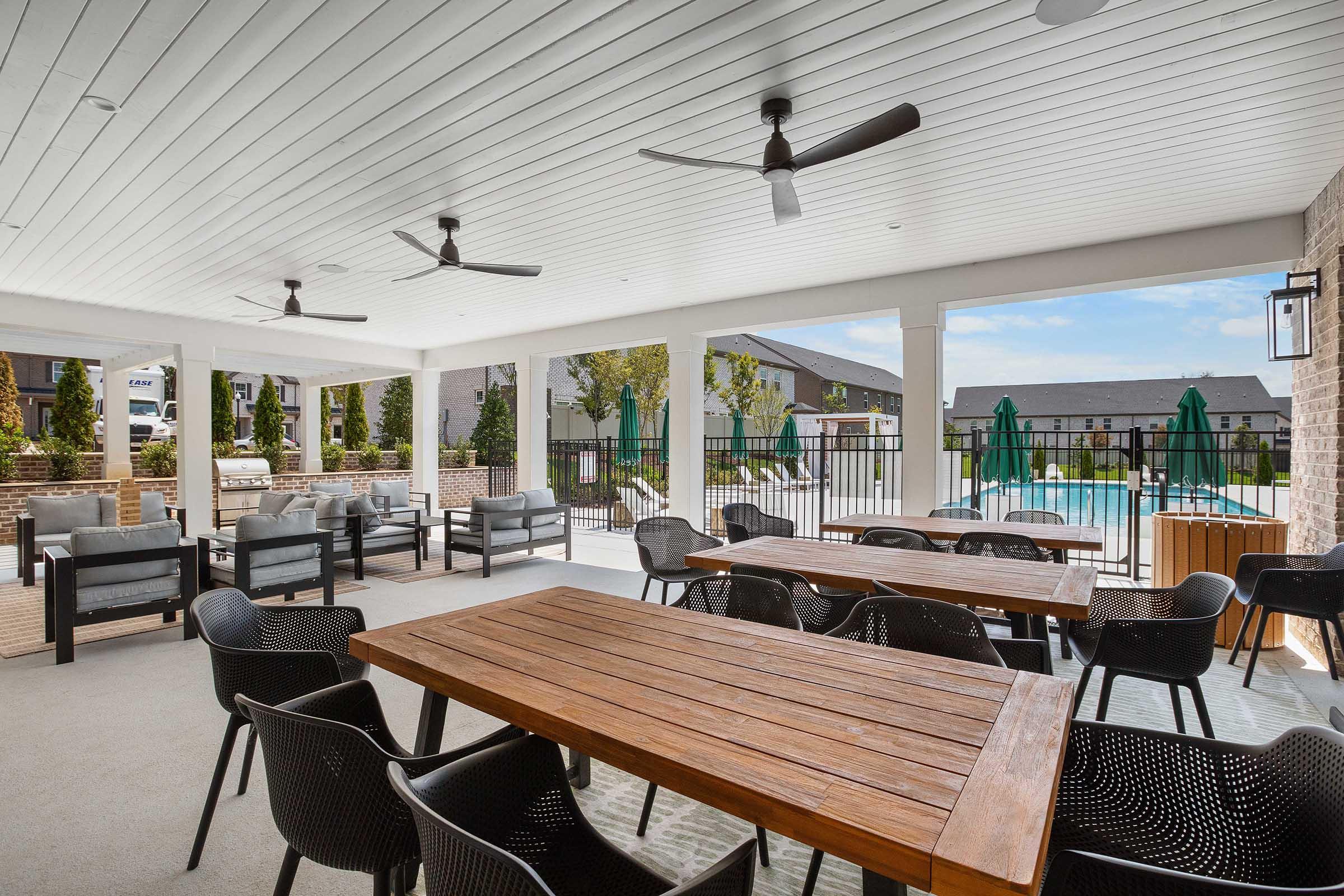 Covered outdoor seating area featuring a large wooden table with black chairs, ceiling fans, and a view of a swimming pool in the background. Lush greenery and patio umbrellas enhance the inviting atmosphere. Ideal for social gatherings or leisure activities.