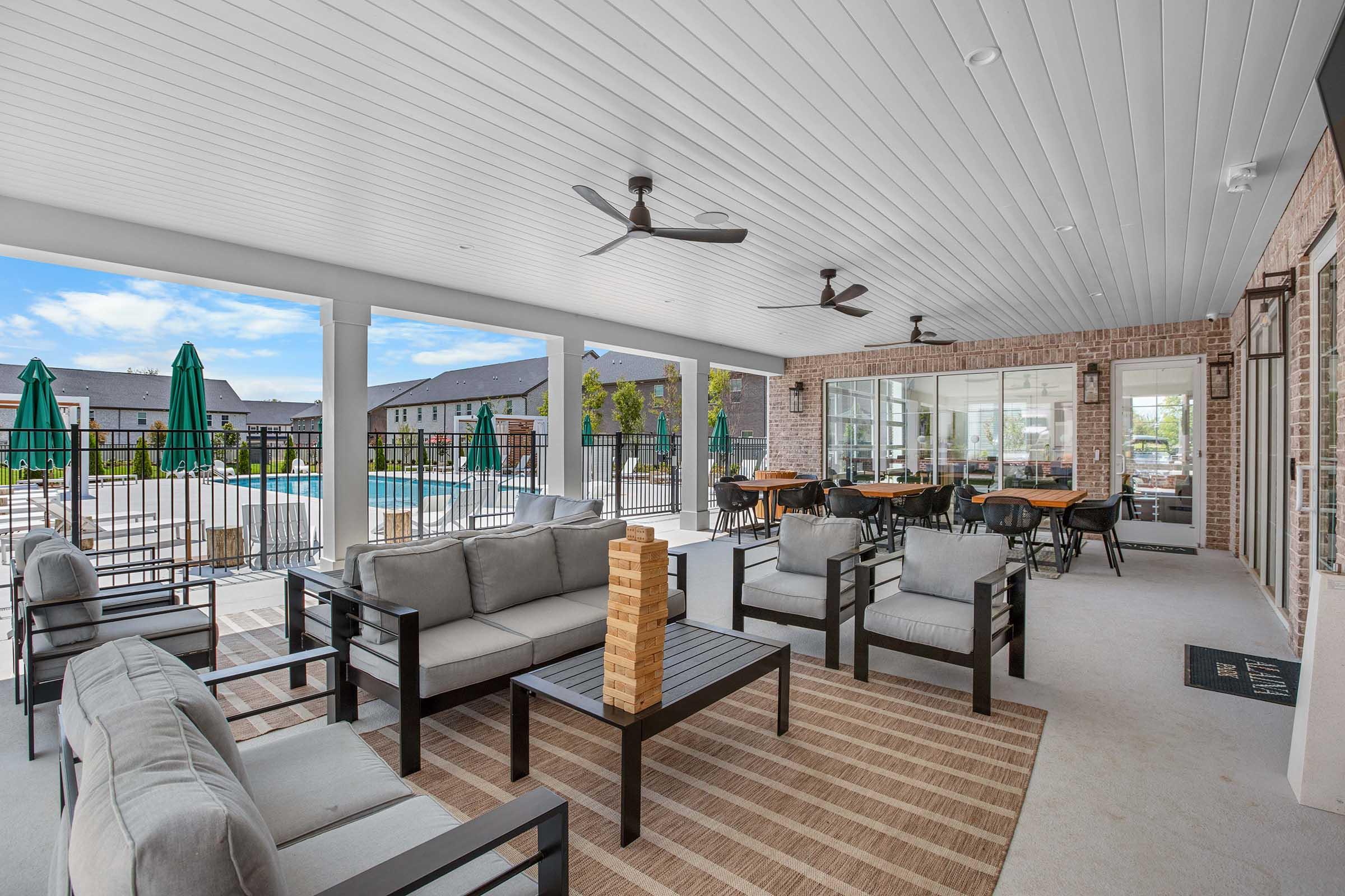 Covered patio area featuring gray outdoor furniture, including sofas and a coffee table with a large wooden game. In the background, there are green umbrellas, a swimming pool, and large windows revealing an indoor dining space. The ceiling has fans for ventilation, and the aesthetic is modern and inviting.