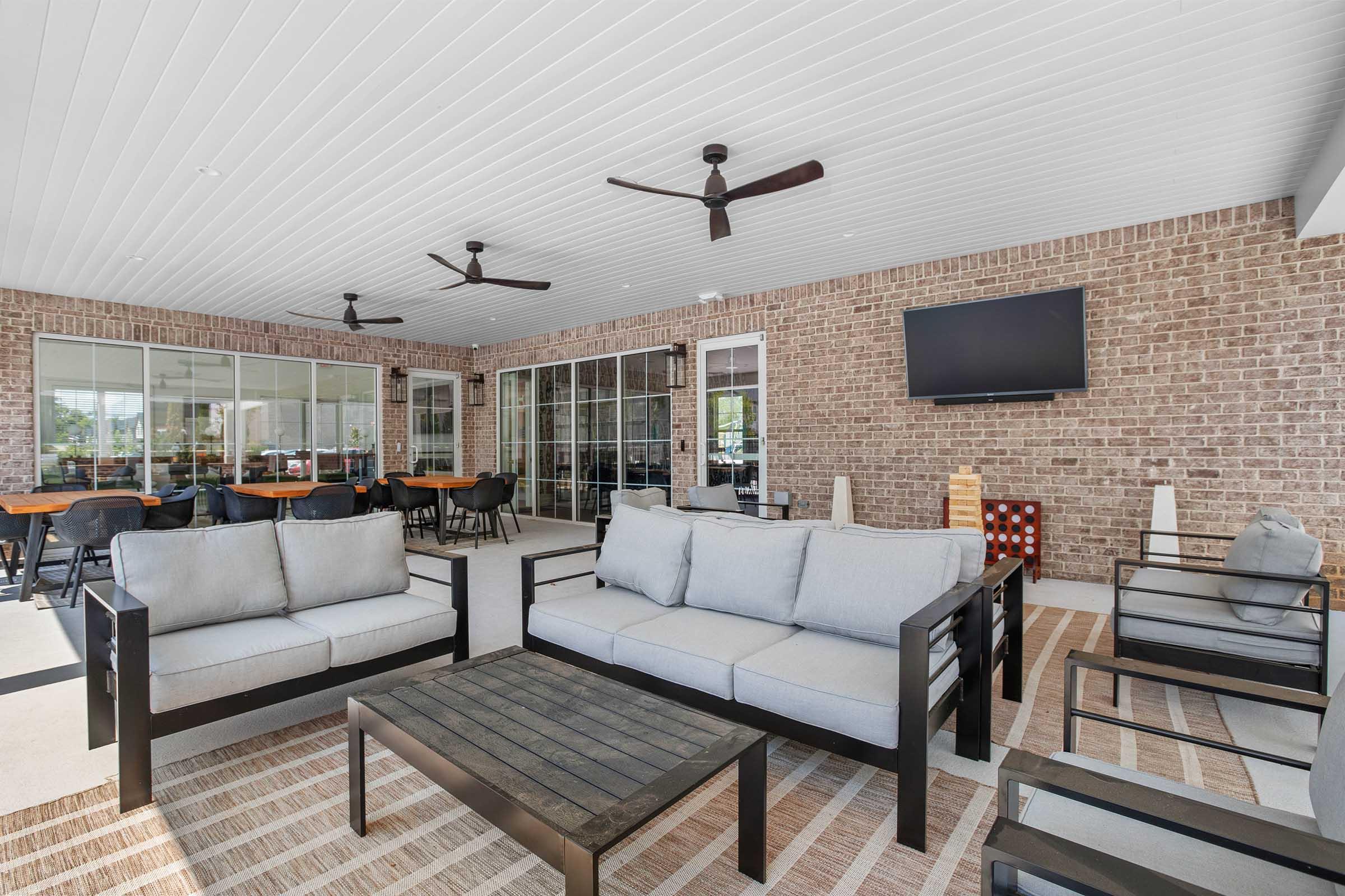 A spacious outdoor lounge area featuring gray sofas with dark wooden frames, a black table, and a modern rug. The area is covered and has ceiling fans, with large glass windows in the background revealing additional seating. A wall-mounted TV is also visible.