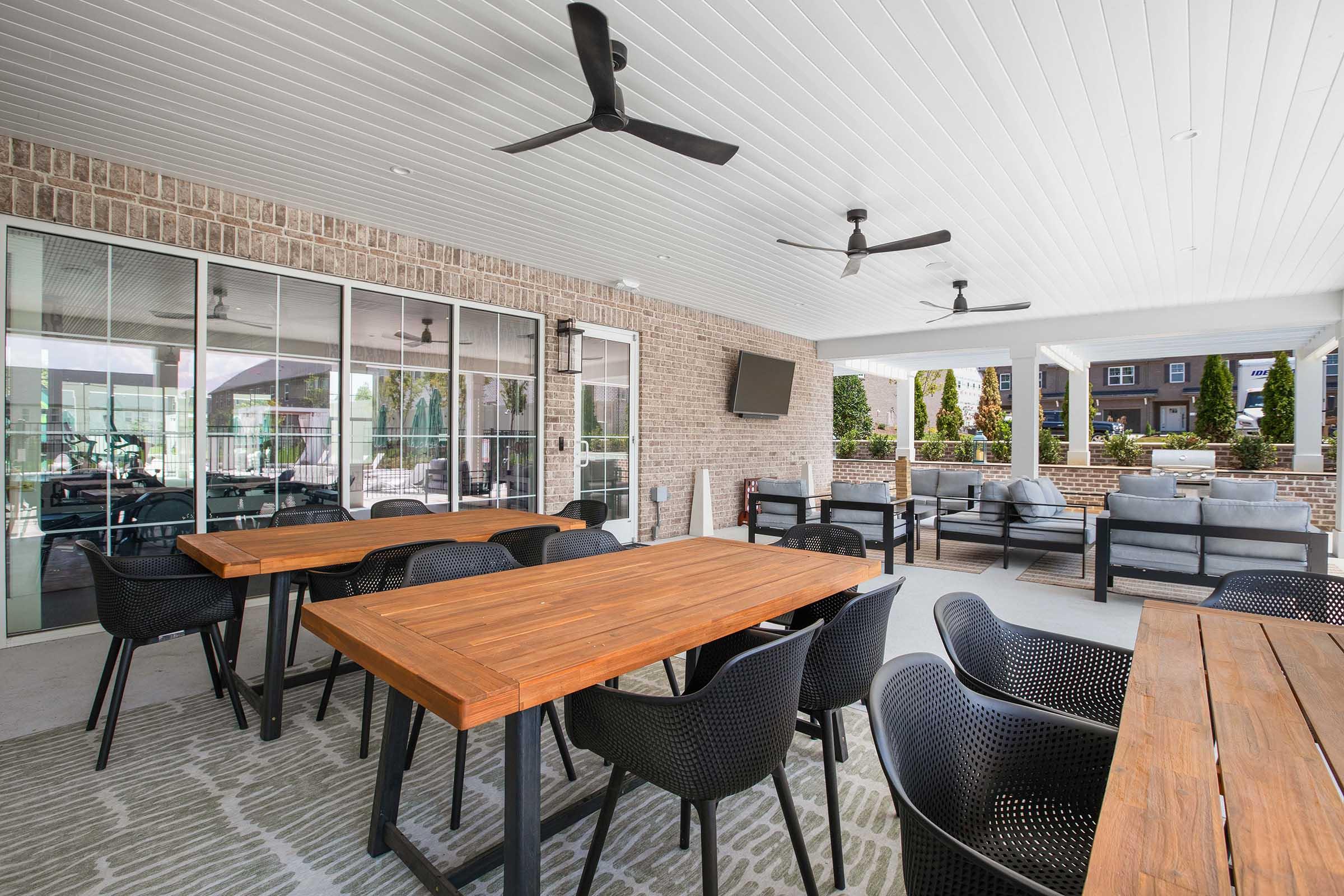 Outdoor patio area featuring wooden tables and black chairs, with ceiling fans for ventilation. The space is partially enclosed with brick walls, includes a television on the wall, and displays comfortable seating arrangements amidst greenery. Ideal for gatherings or relaxation.