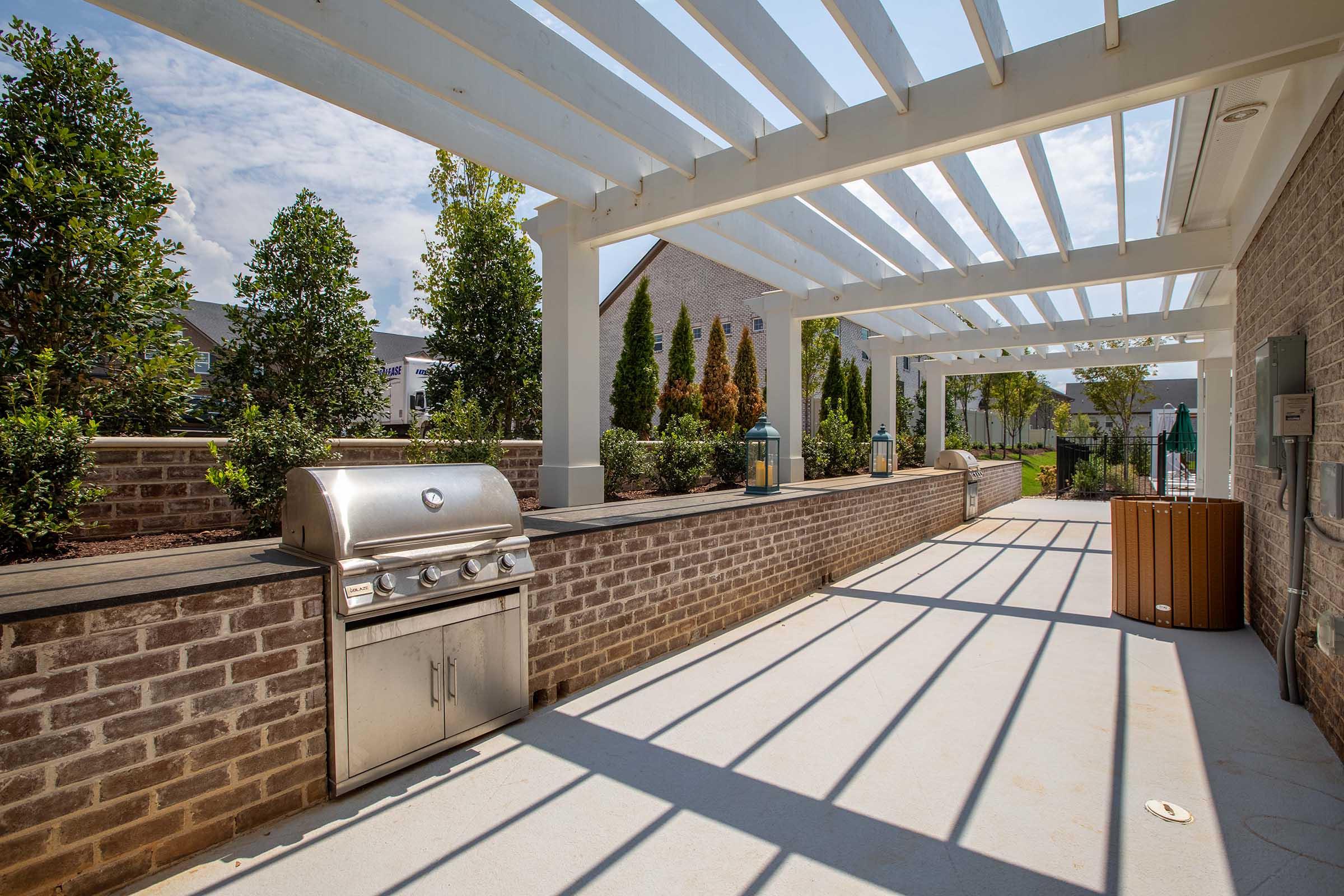 Outdoor patio space with a built-in stainless steel grill, surrounded by brick walls and landscaped greenery. The area features a pergola overhead, allowing sunlight to create distinct shadows on the concrete floor, with seating areas and decorative plants in the background.