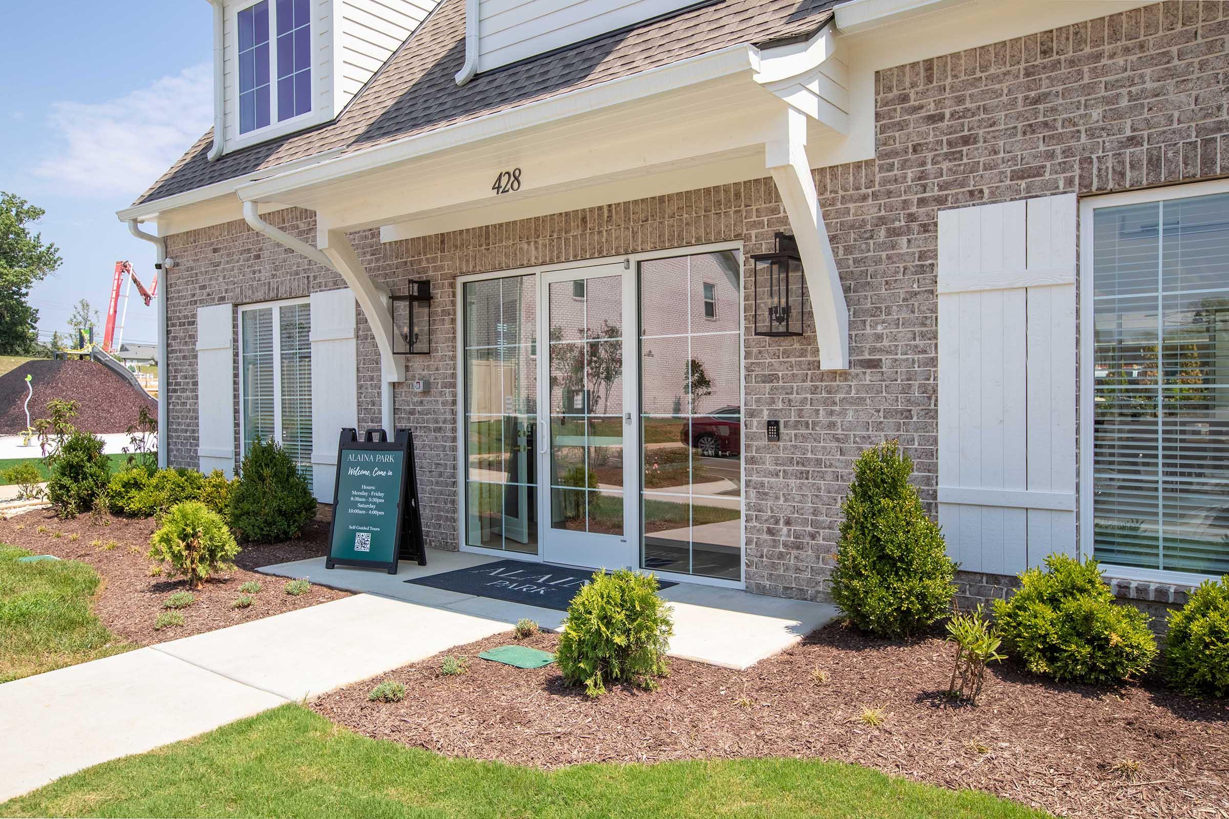 Entrance of a brick building featuring large glass doors with a sign beside it that displays the number "428." The area is landscaped with small shrubs and has a clear walkway. Sunlight illuminates the scene, creating a welcoming atmosphere.