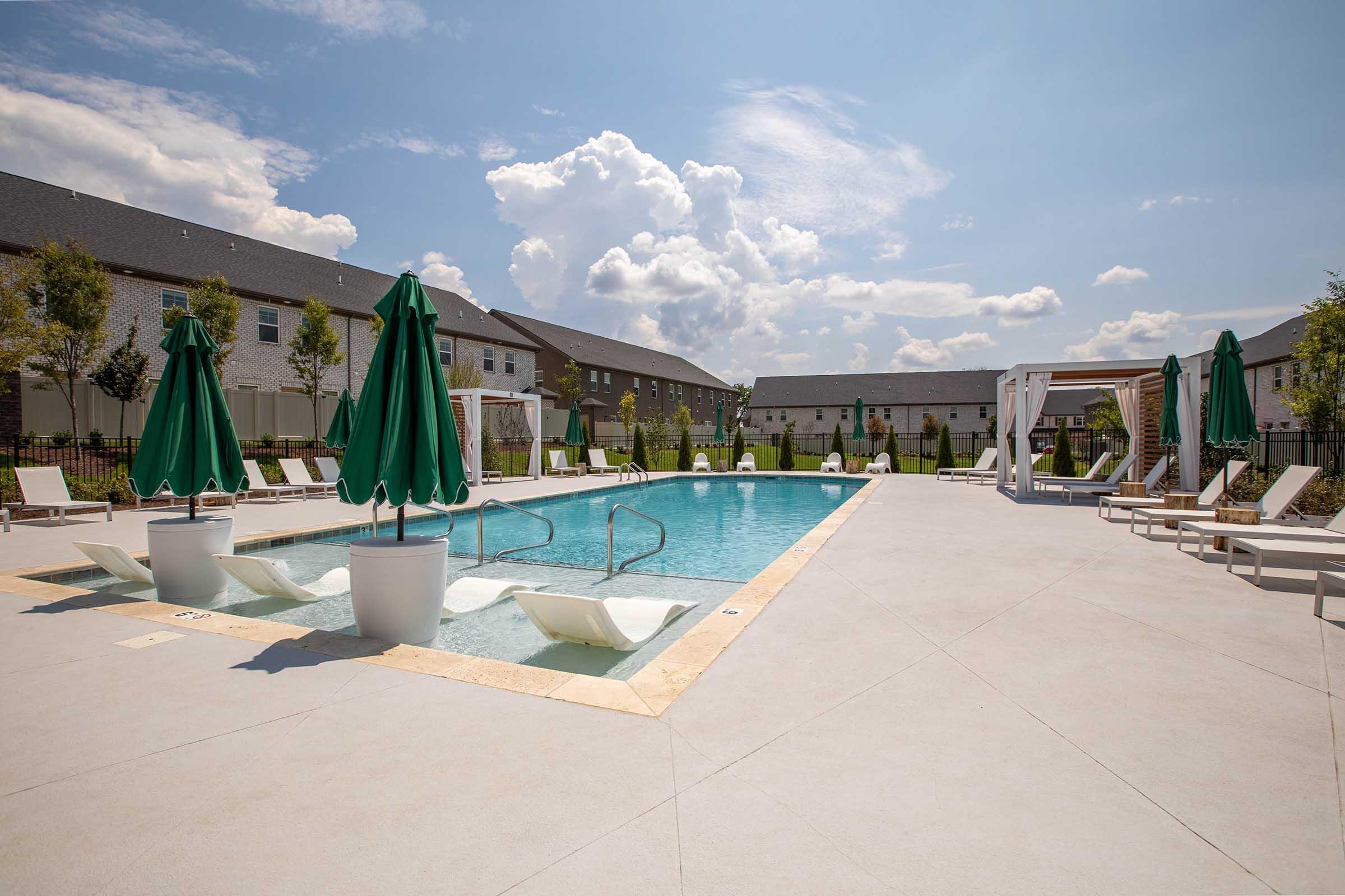 A sunny outdoor pool area with a clear blue water pool surrounded by white lounge chairs and green umbrellas. Lush landscaping is visible in the background, along with several shade structures. The sky is mostly cloudy, creating a vibrant atmosphere for relaxation and enjoyment.