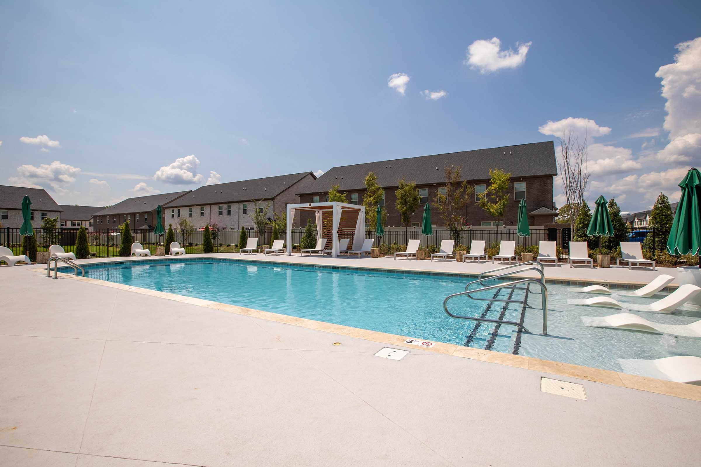 A bright outdoor swimming pool surrounded by lounge chairs and green umbrellas, with several trees in the background. The sky is clear with a few clouds, and there are residential buildings nearby. The pool area is well-maintained and inviting for relaxation.