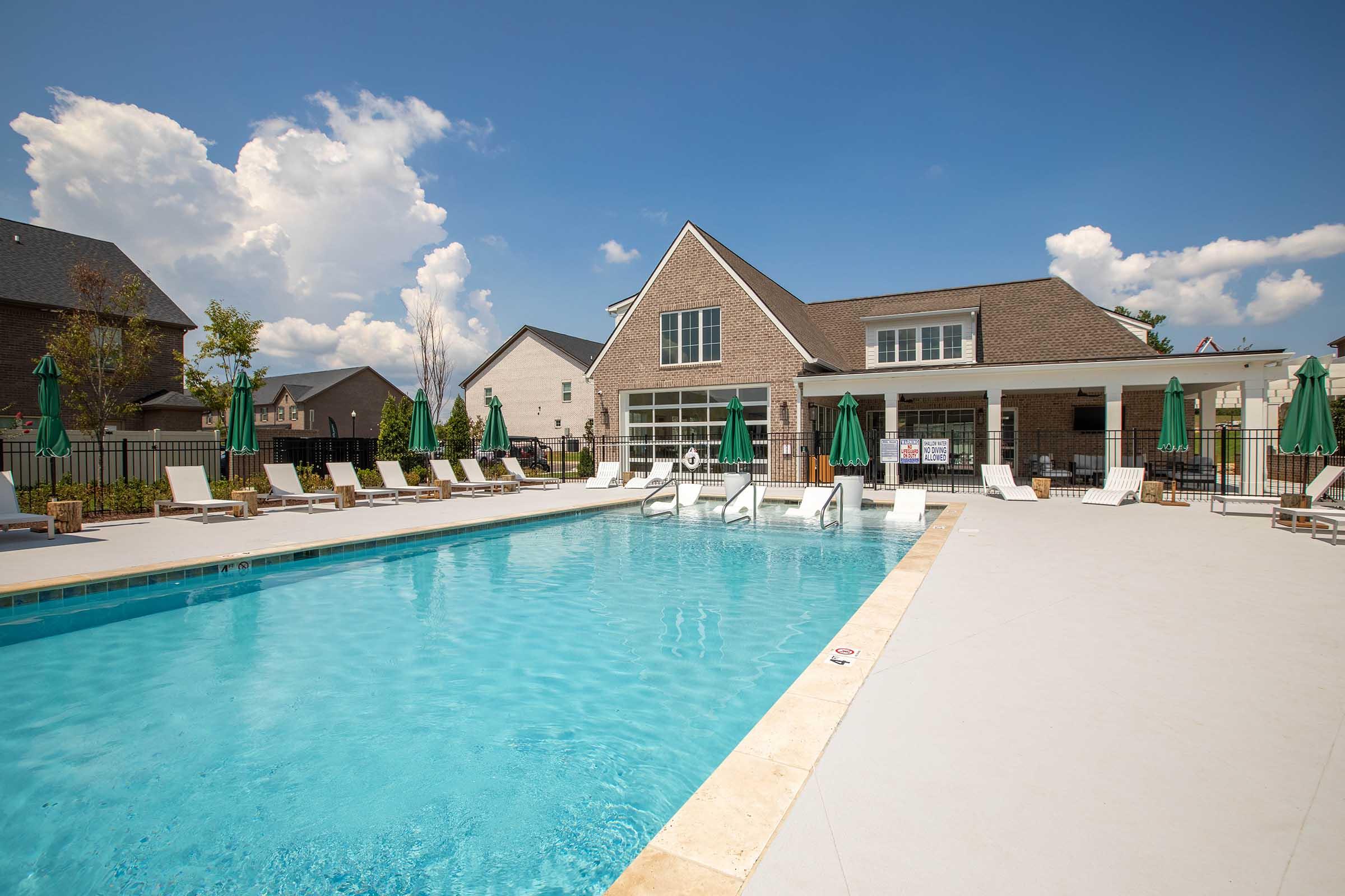 A bright and inviting swimming pool surrounded by lounge chairs and green umbrellas. The pool area features a modern building in the background with large windows, under a clear blue sky with fluffy white clouds, creating a relaxing atmosphere for leisure and enjoyment.